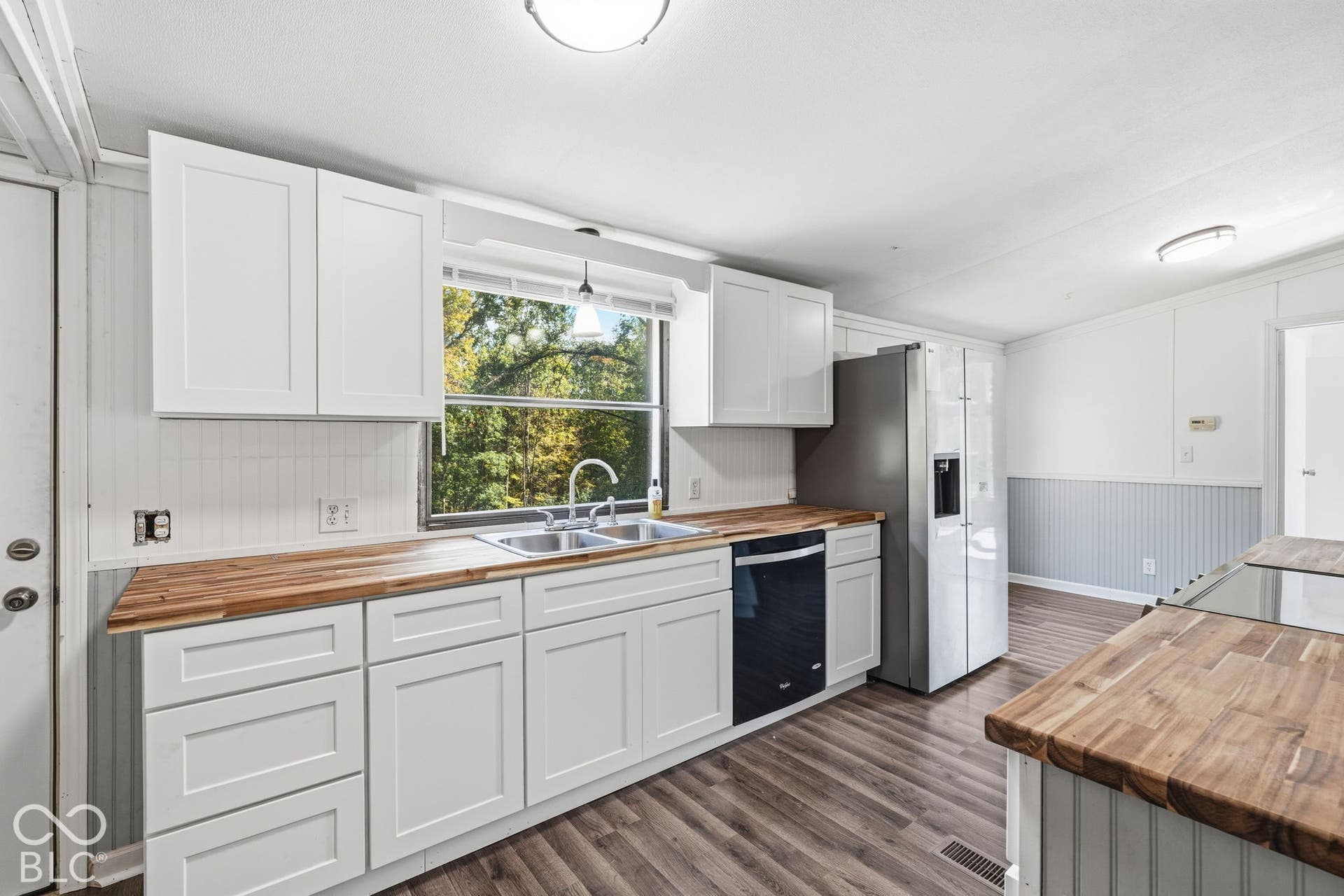 Kitchen featuring white shaker-style cabinetry, wood-finish countertops, dual-basin stainless steel sink, beadboard wainscoting, and wood-finish flooring