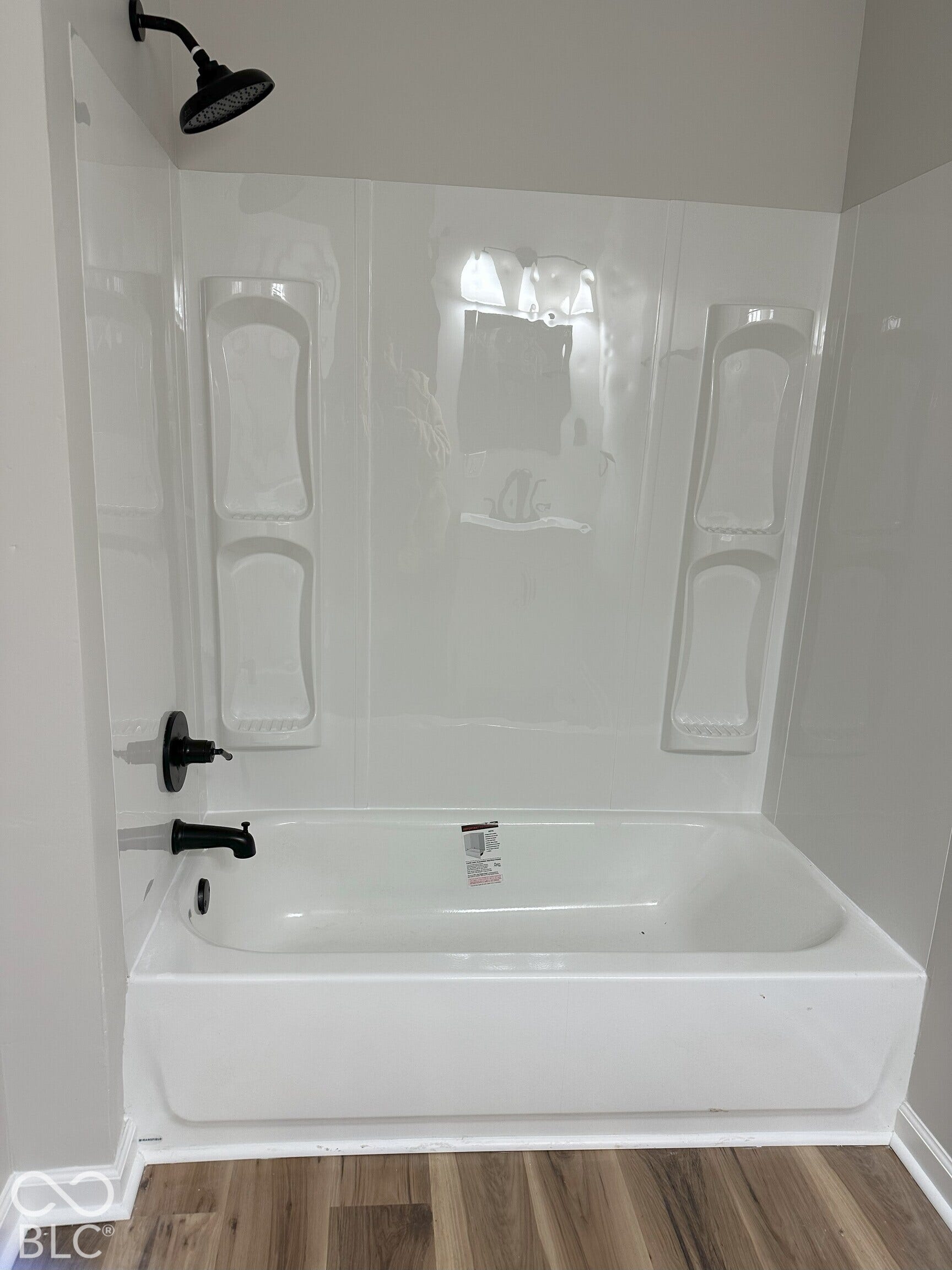 Bathtub enclosure featuring integrated molded shelving, matte black wall-mounted showerhead, matte black faucet hardware, and wood-finish flooring