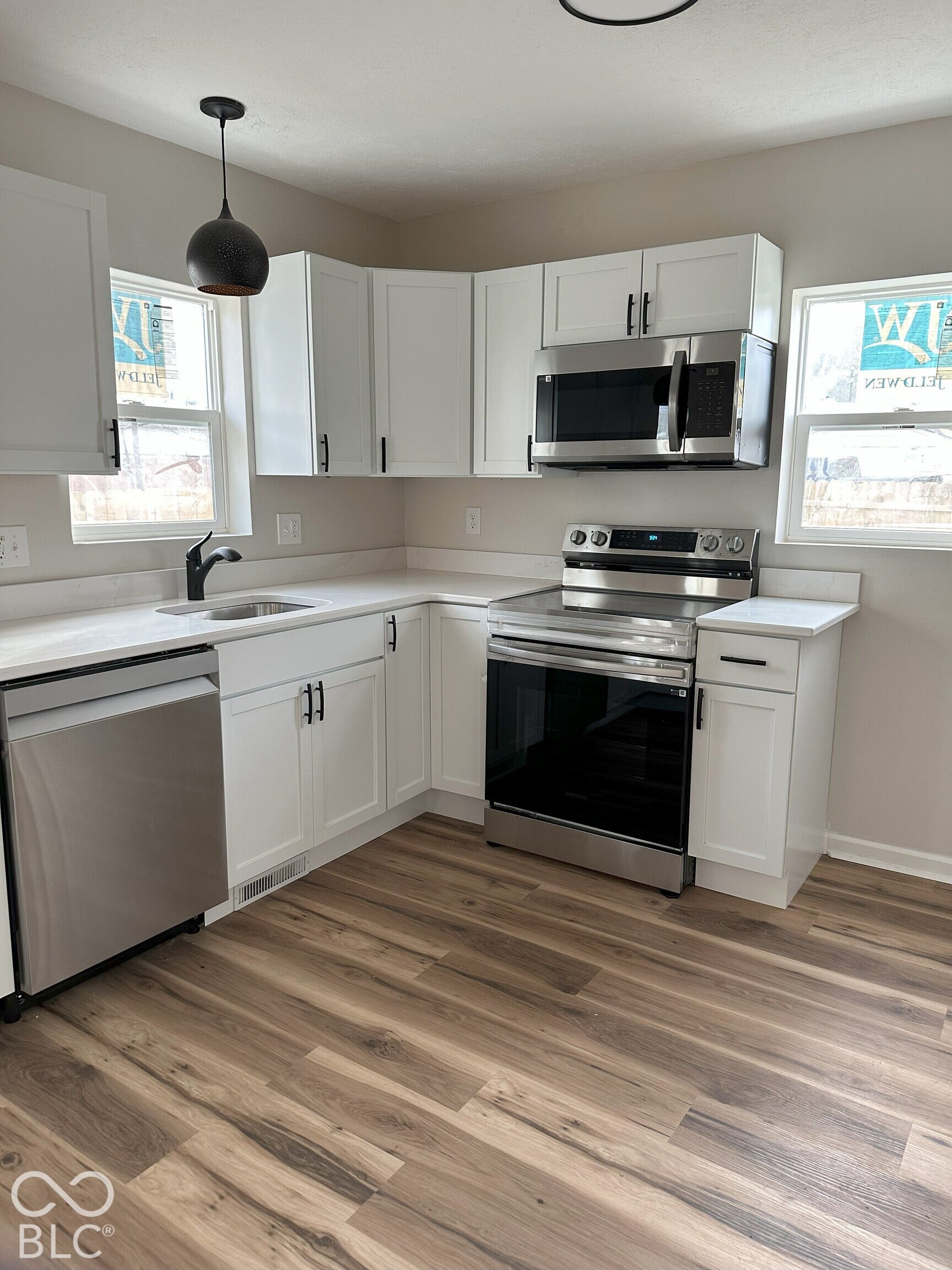 Kitchen featuring white shaker-style cabinetry, stainless steel appliances, wood-finish flooring, white solid-surface countertops, and a matte black pendant light fixture
