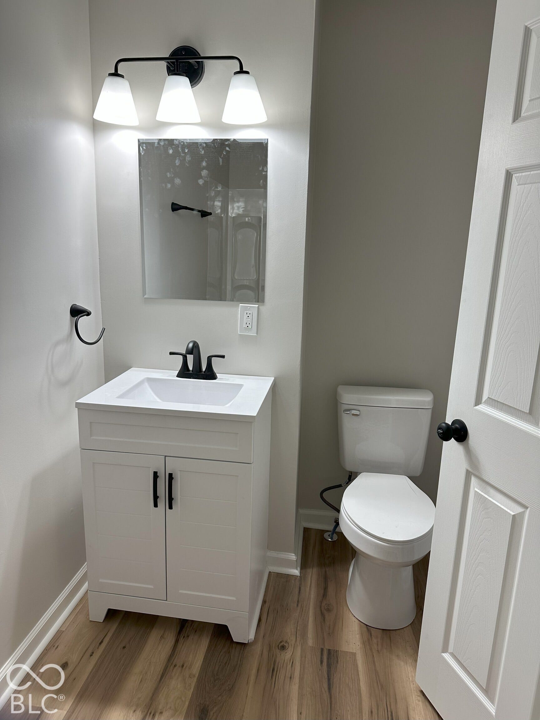 White cabinetry vanity with a clean-lined rectangular sink, matte black faucet, coordinating three-light vanity fixture, and durable wood-finish flooring