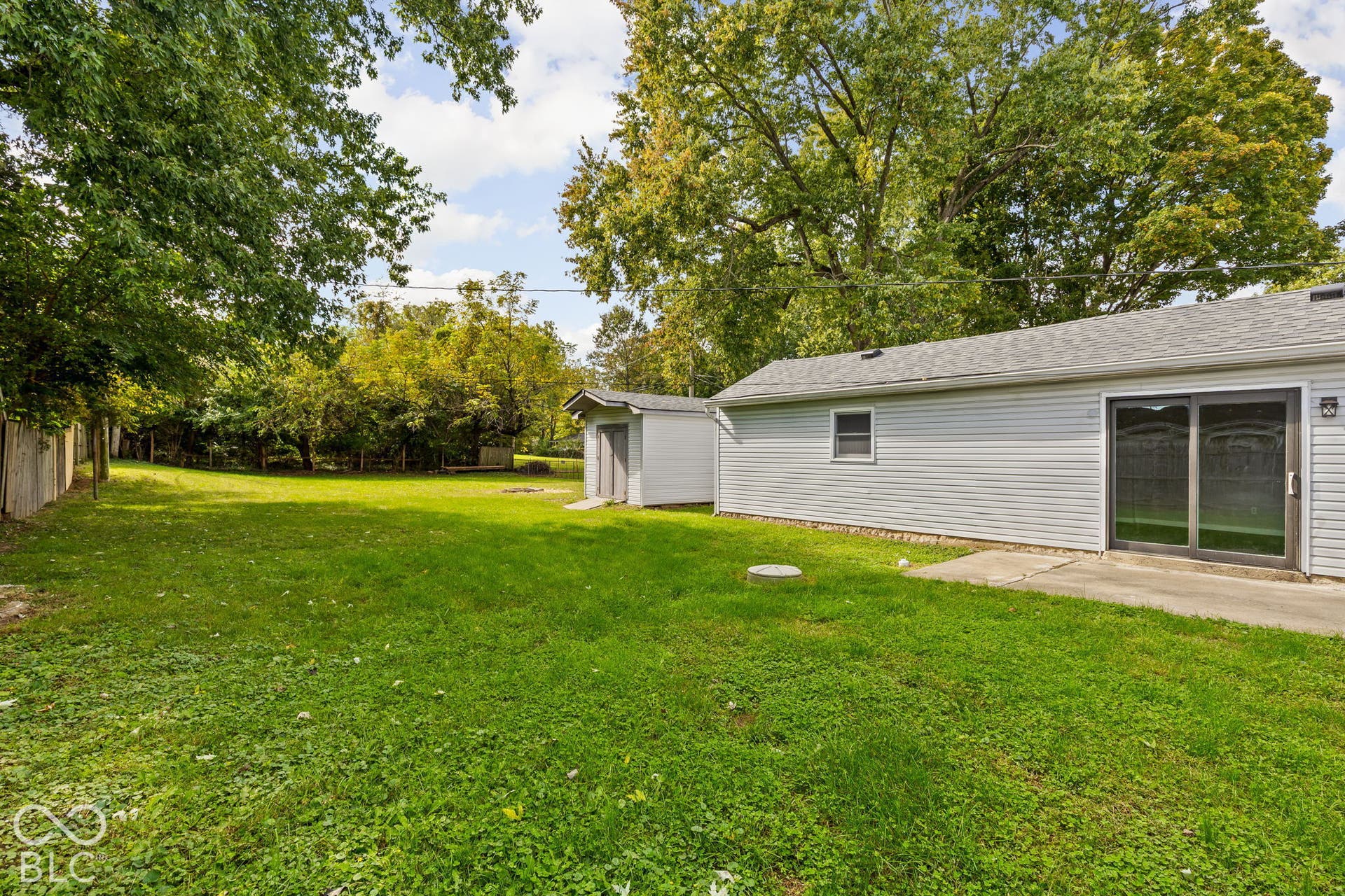Expansive backyard featuring a concrete patio, detached storage shed, mature trees, and perimeter wood fencing