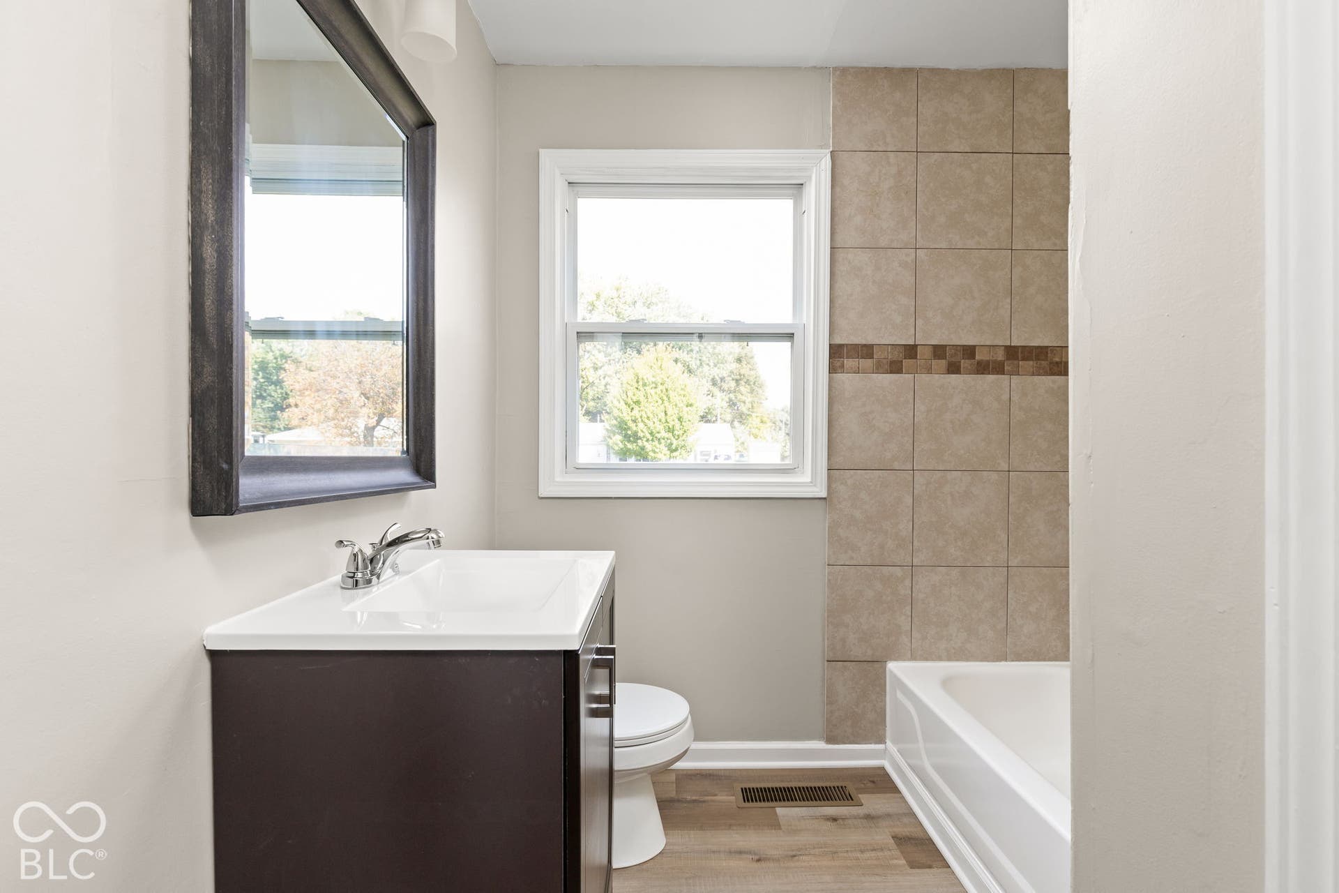 Bathroom featuring a dark wood-finish vanity, integrated white sink, wall-mounted mirror, wood-finish flooring, and a tub enclosure with neutral ceramic wall tiling
