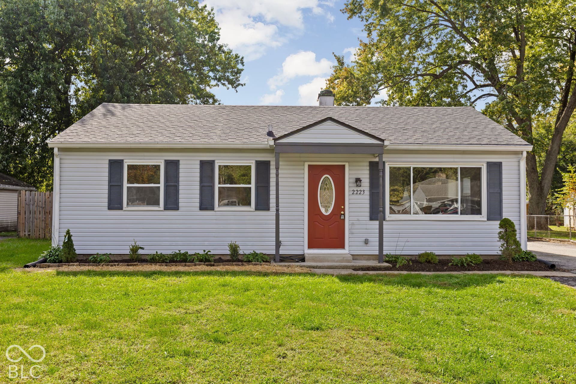 Single-story exterior featuring light-toned horizontal siding, dark decorative shutters, an accented red front door with an oval glass insert, and a covered entryway with a peaked roofline