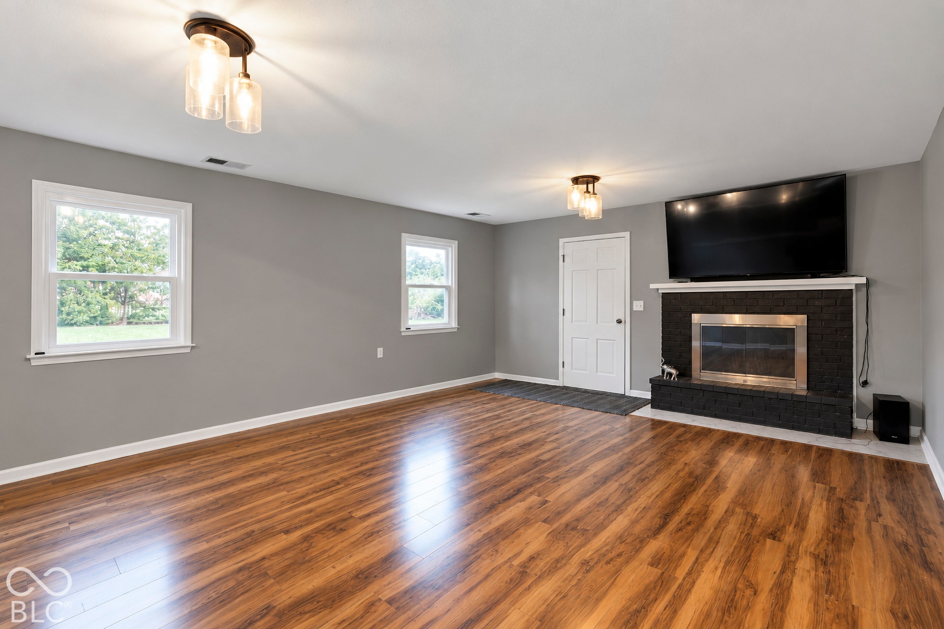 Living area featuring wood-finish flooring, a fireplace with a black brick surround, two multi-bulb flush-mount light fixtures, and windows with white trim
