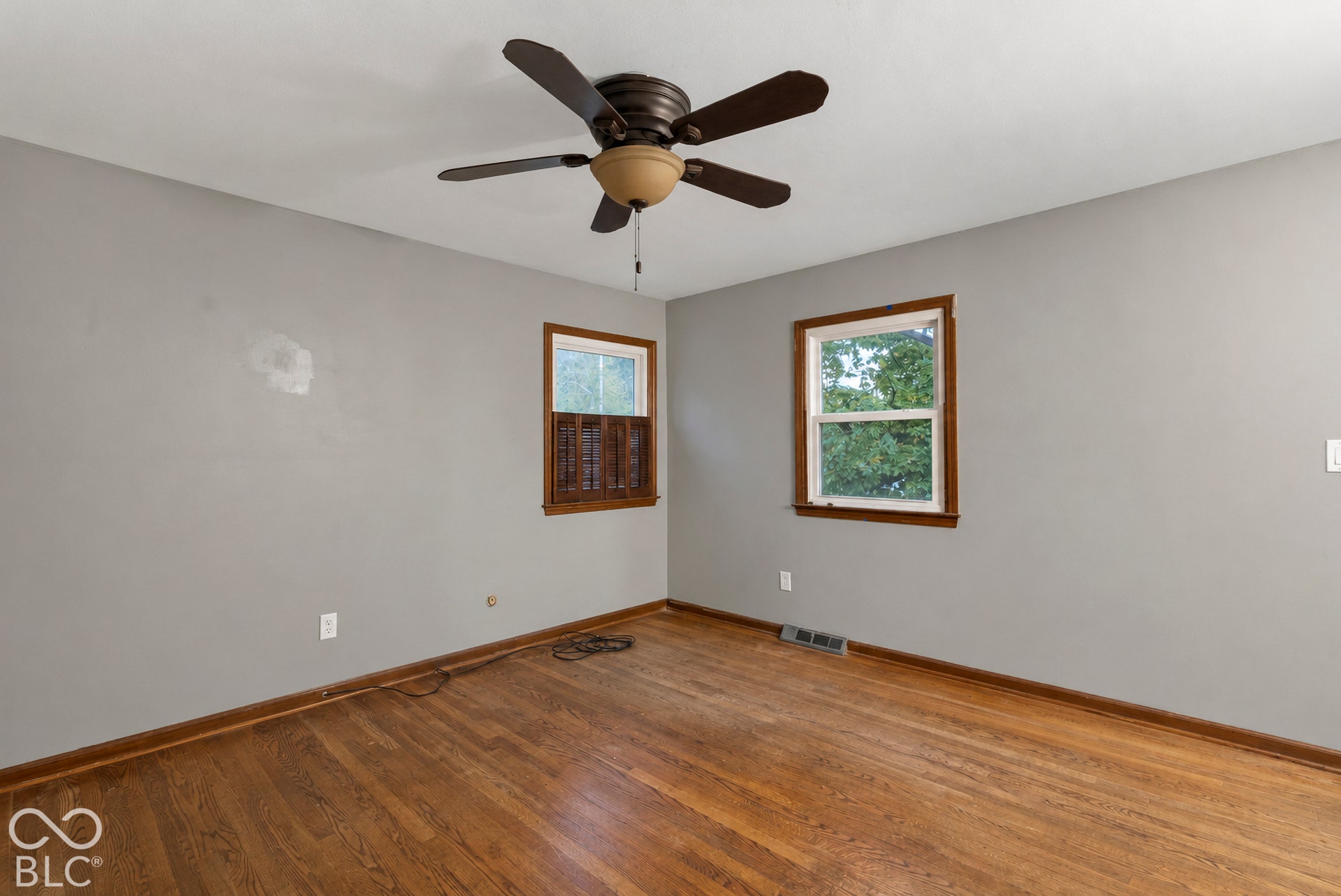 Wood-finish flooring, neutral wall paint, two windows with wood-tone casing, and a multi-blade ceiling fan with central lighting