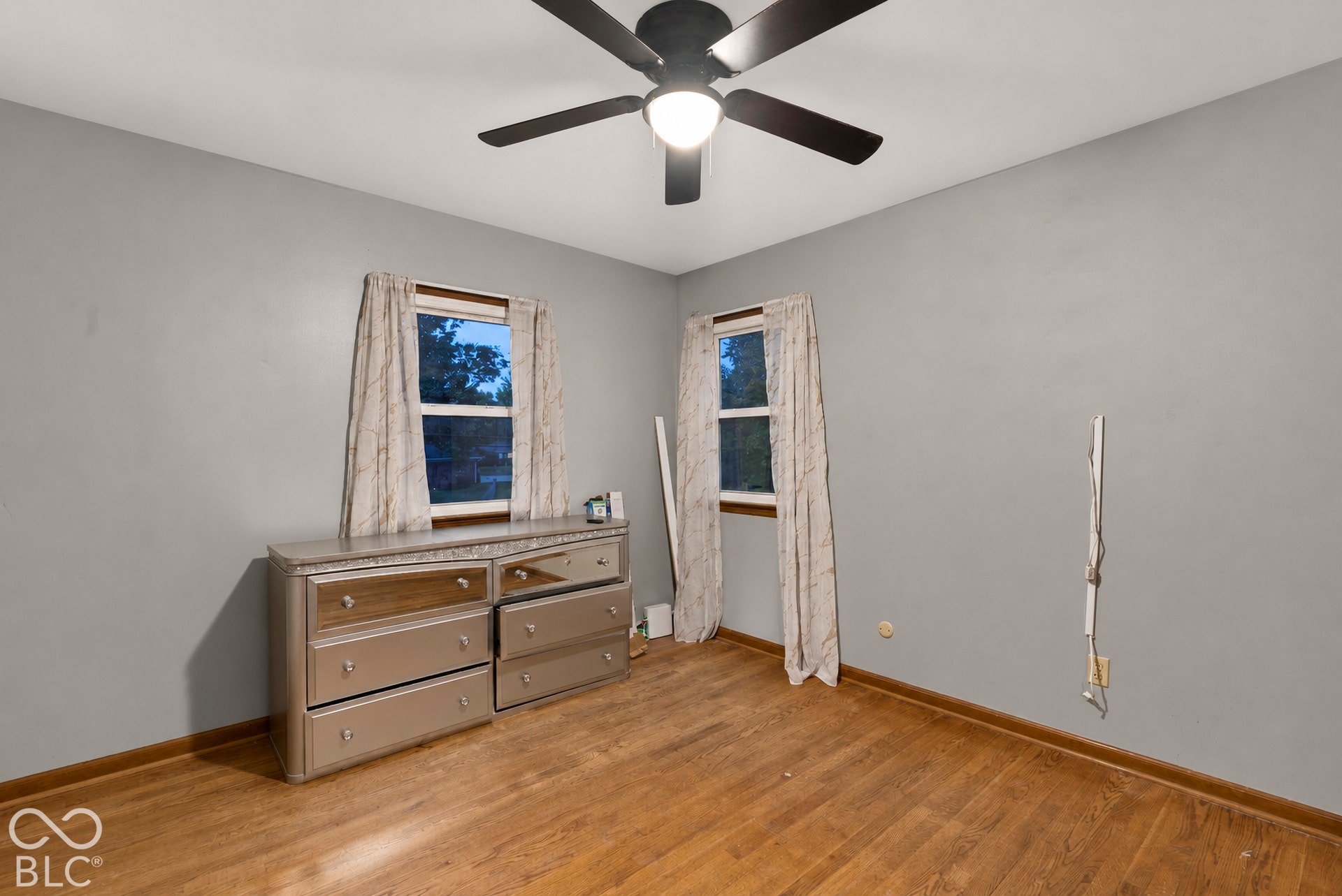 Room interior featuring wood-finish flooring, two dual-pane windows with trim, and a ceiling-mounted matte black light fixture with a fan
