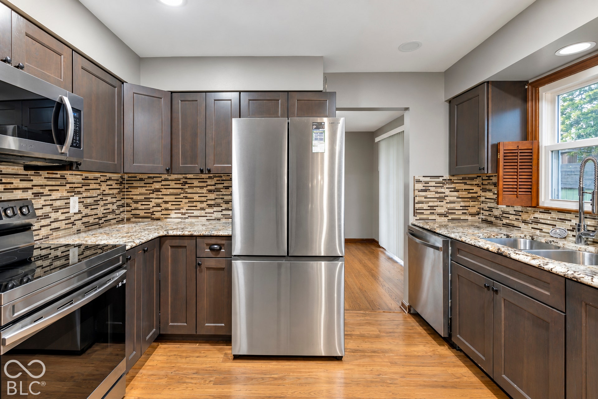 Kitchen featuring shaker-style cabinetry, stone countertops, integrated stainless steel appliances, multi-tonal tile backsplash, and wood-finish flooring