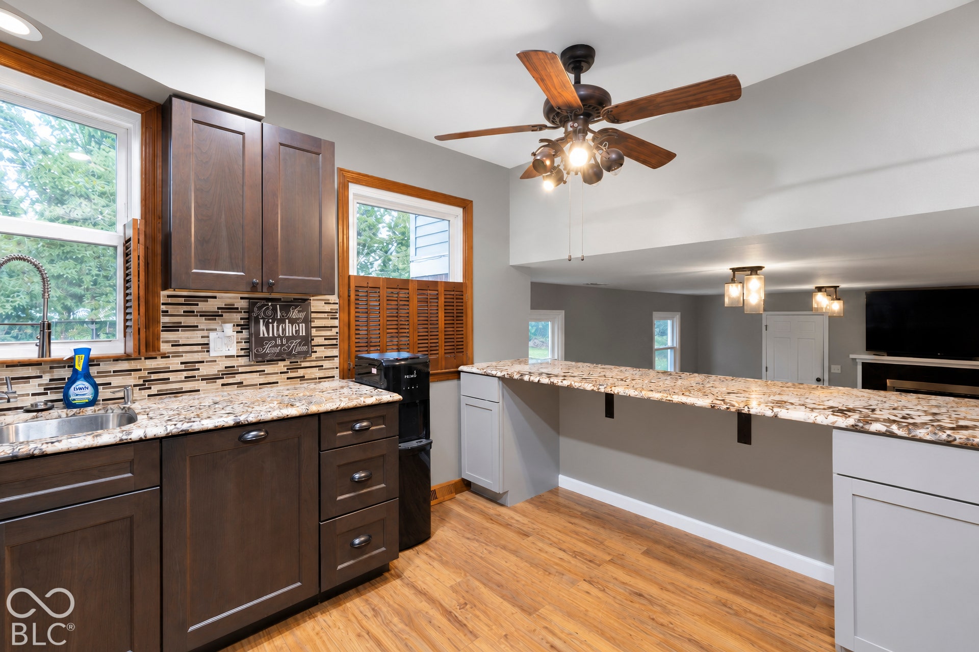 Kitchen featuring dark cabinetry, stone-finish countertops, decorative tile backsplash, wood-finish flooring, and a ceiling fan with multi-bulb light fixture