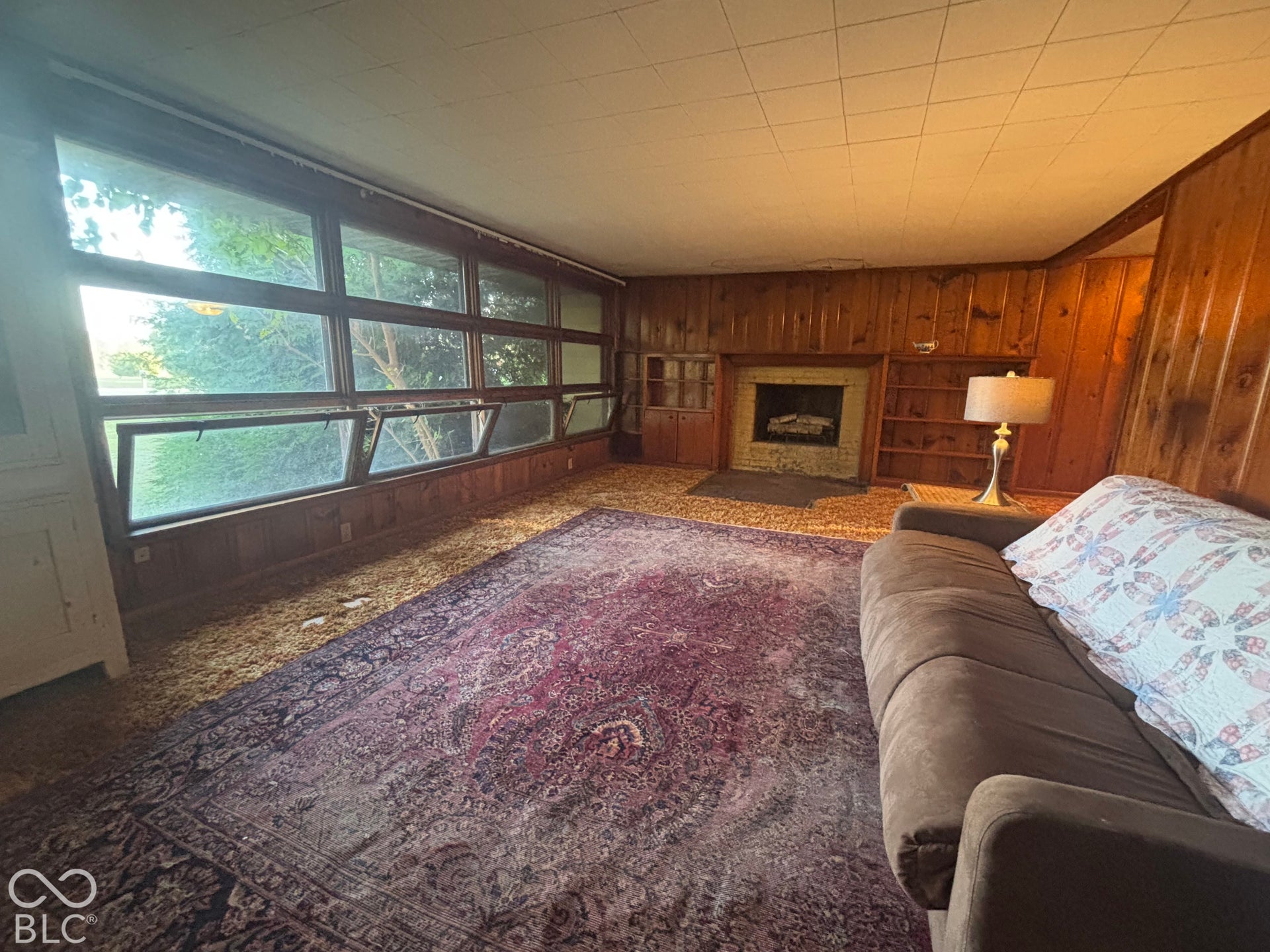 Living area featuring wood-paneled walls, large multi-pane windows with hopper-style openings, a built-in fireplace, and integrated wooden shelving