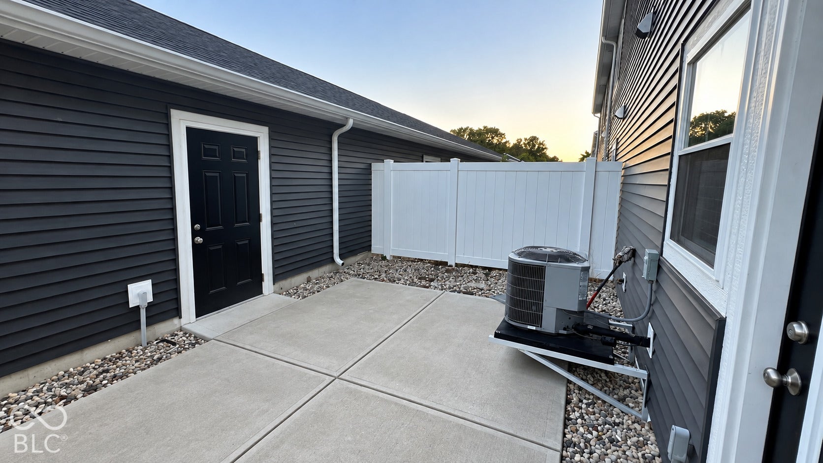 Dark horizontal lap siding exterior featuring a concrete patio, white vinyl privacy fencing, and exterior entry doors