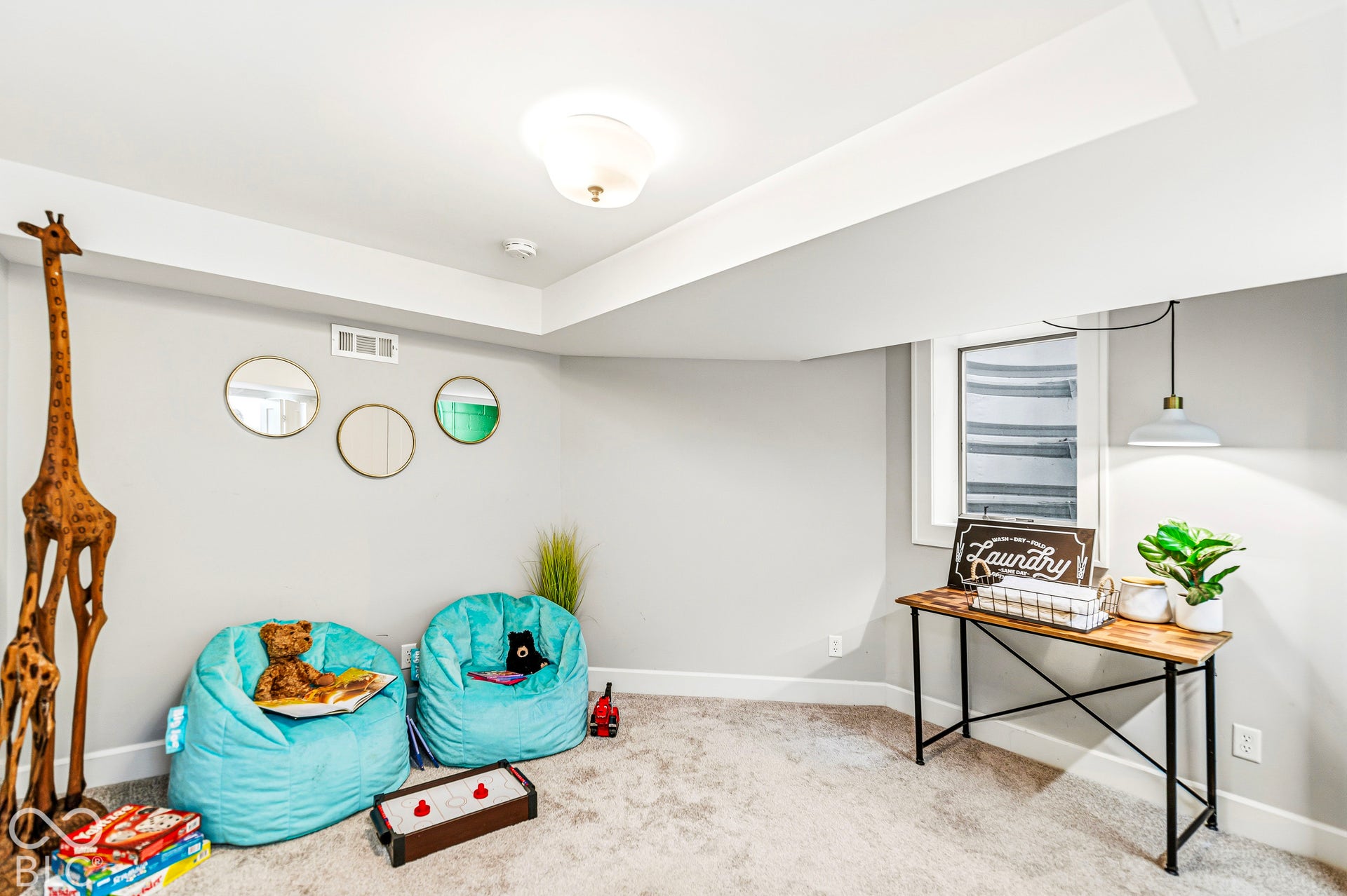 Neutral-toned interior space featuring wall-to-wall carpeting, an egress window with a window well, a flush-mount ceiling light fixture, and a pendant light