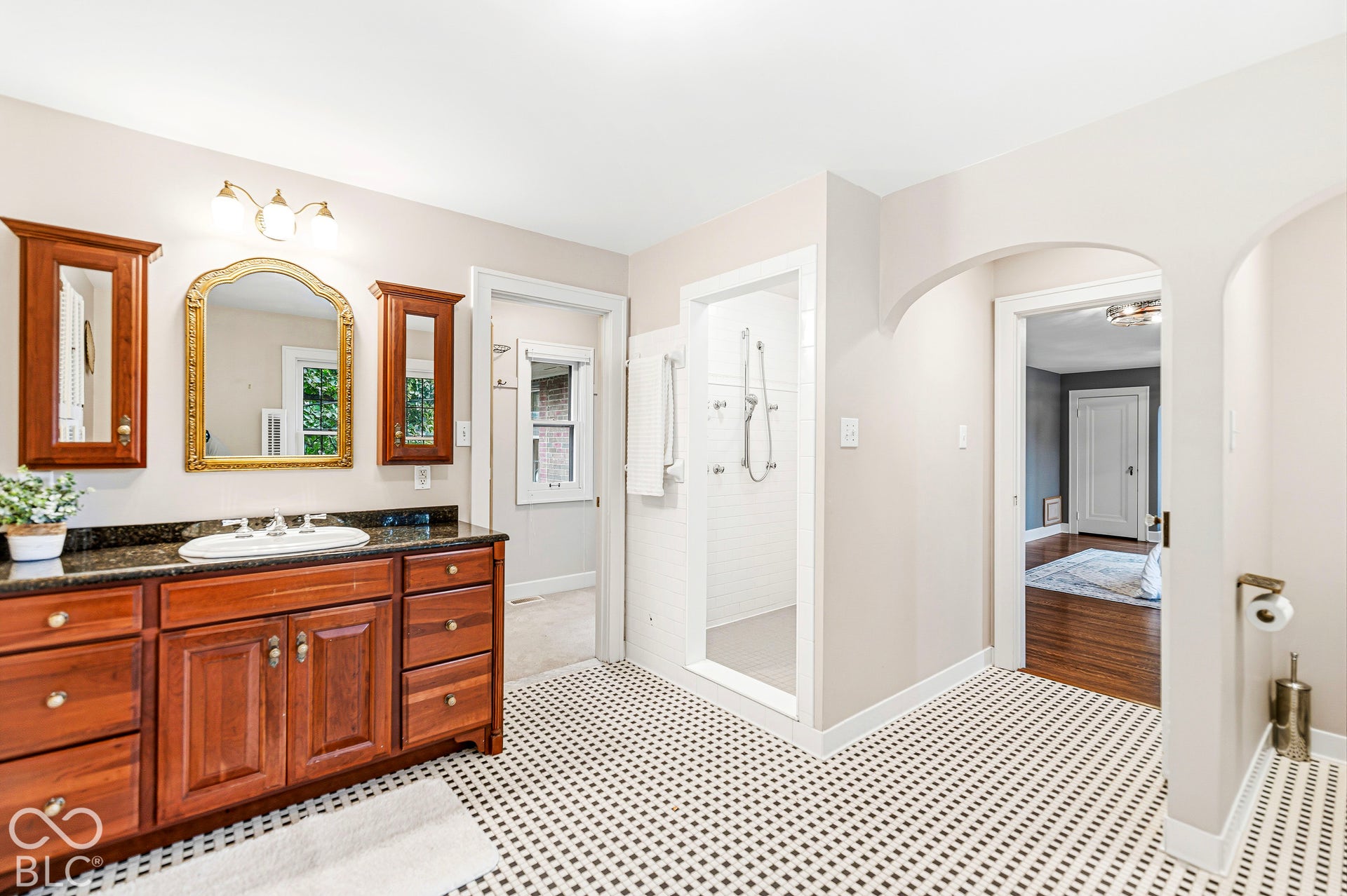 Custom wood-finish cabinetry, dark stone vanity countertop, arched doorway, walk-in shower with white tile surround, and black-and-white mosaic tile flooring