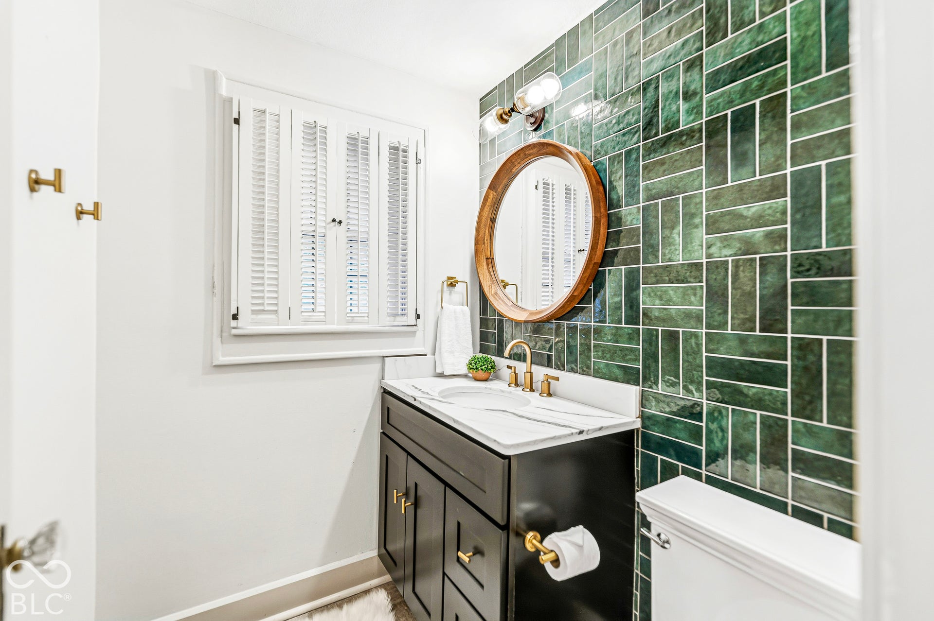 Bathroom featuring an emerald green tiled feature wall, a black vanity with a stone countertop, brass hardware, and white plantation shutters