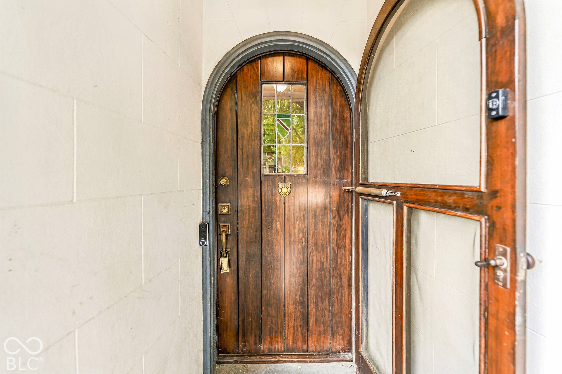 Arched solid wood front door featuring vertical plank construction, decorative leaded glass inlay, brass hardware, and a matching arched wood-frame storm door set within a light-toned masonry entryway