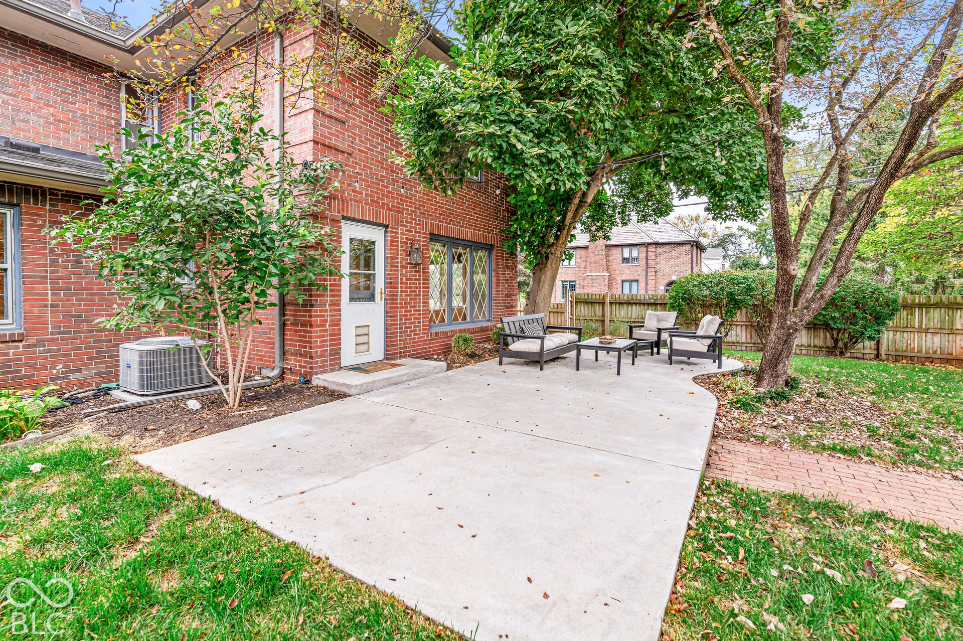 Brick exterior featuring a concrete patio, diamond-pane window detailing, mature trees, and wooden privacy fencing