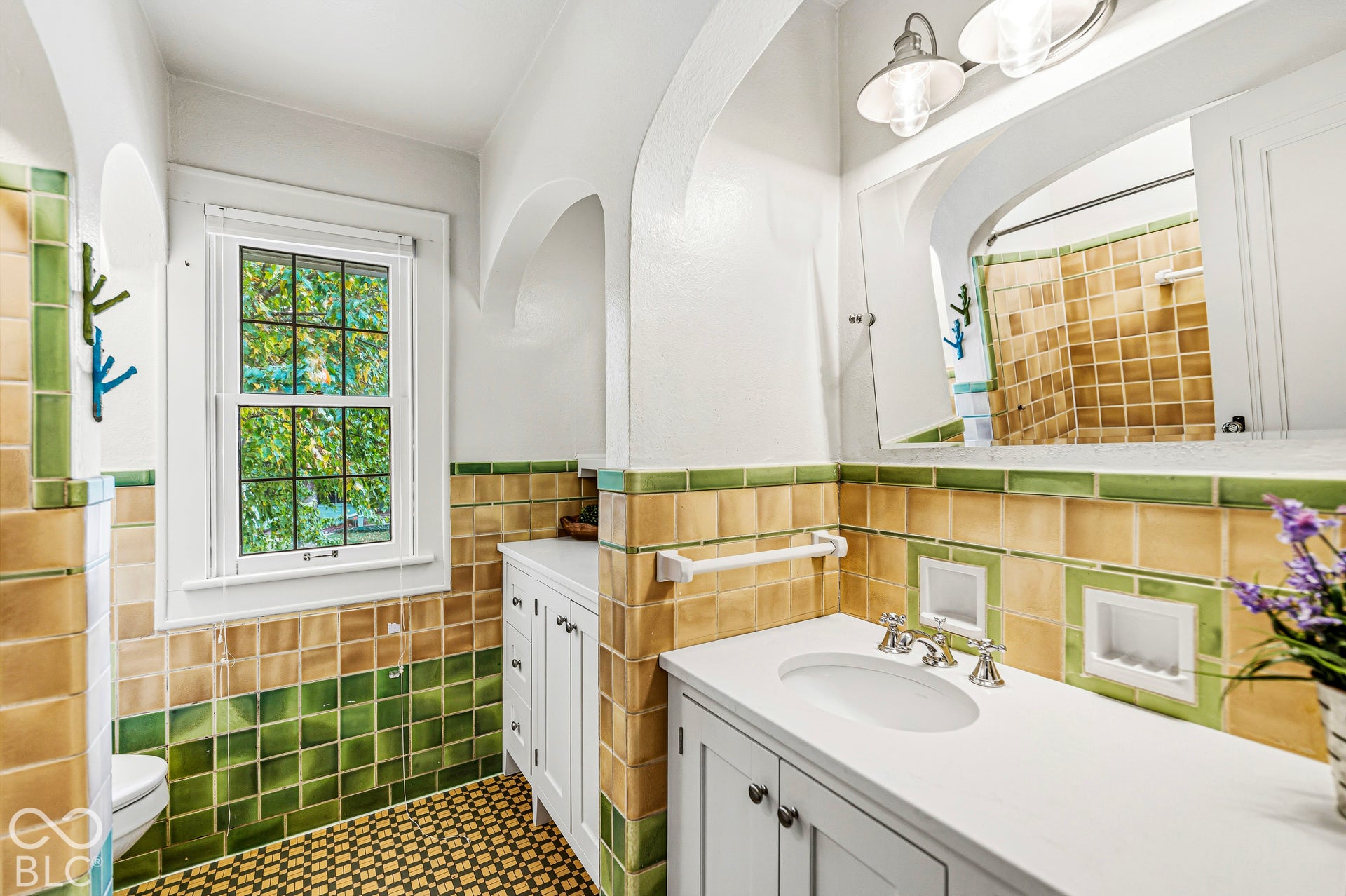 Custom white cabinetry with a light-toned countertop, dual-tone ceramic tile wainscoting, arched architectural detailing, polished metal hardware, and multi-light wall fixtures