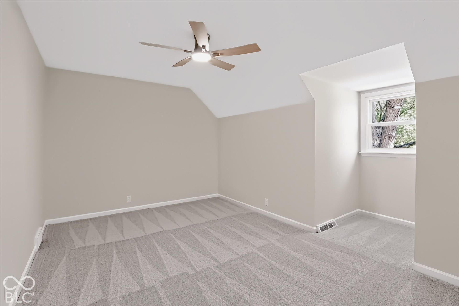 Neutral-toned interior space featuring vaulted ceilings, wall-to-wall carpeting, a recessed window nook, and a ceiling fan with integrated light fixture