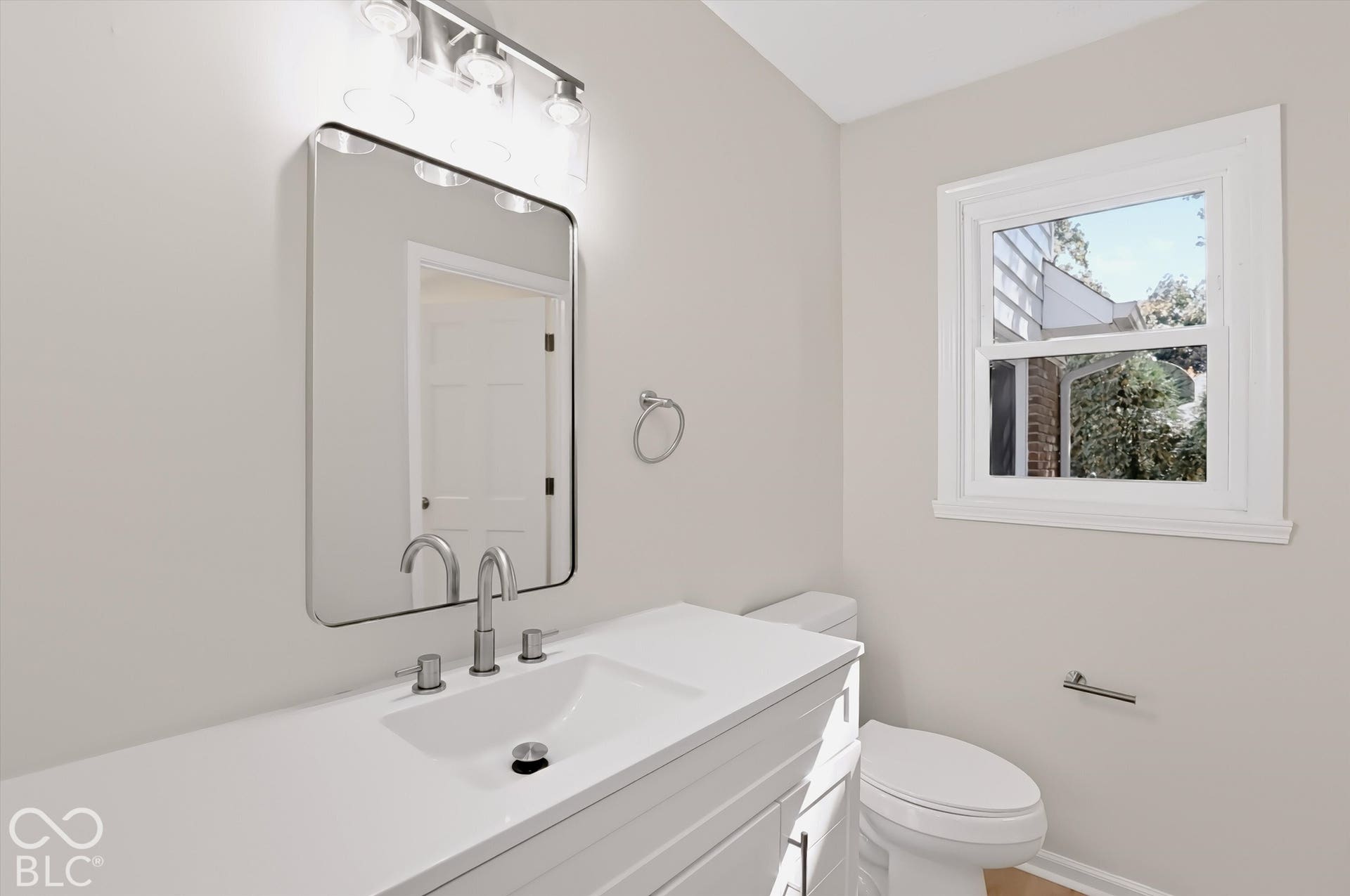 Bathroom featuring a white vanity cabinet with a stone countertop, an integrated rectangular sink with a chrome widespread faucet, a wall-mounted rectangular mirror, and a three-light polished chrome fixture