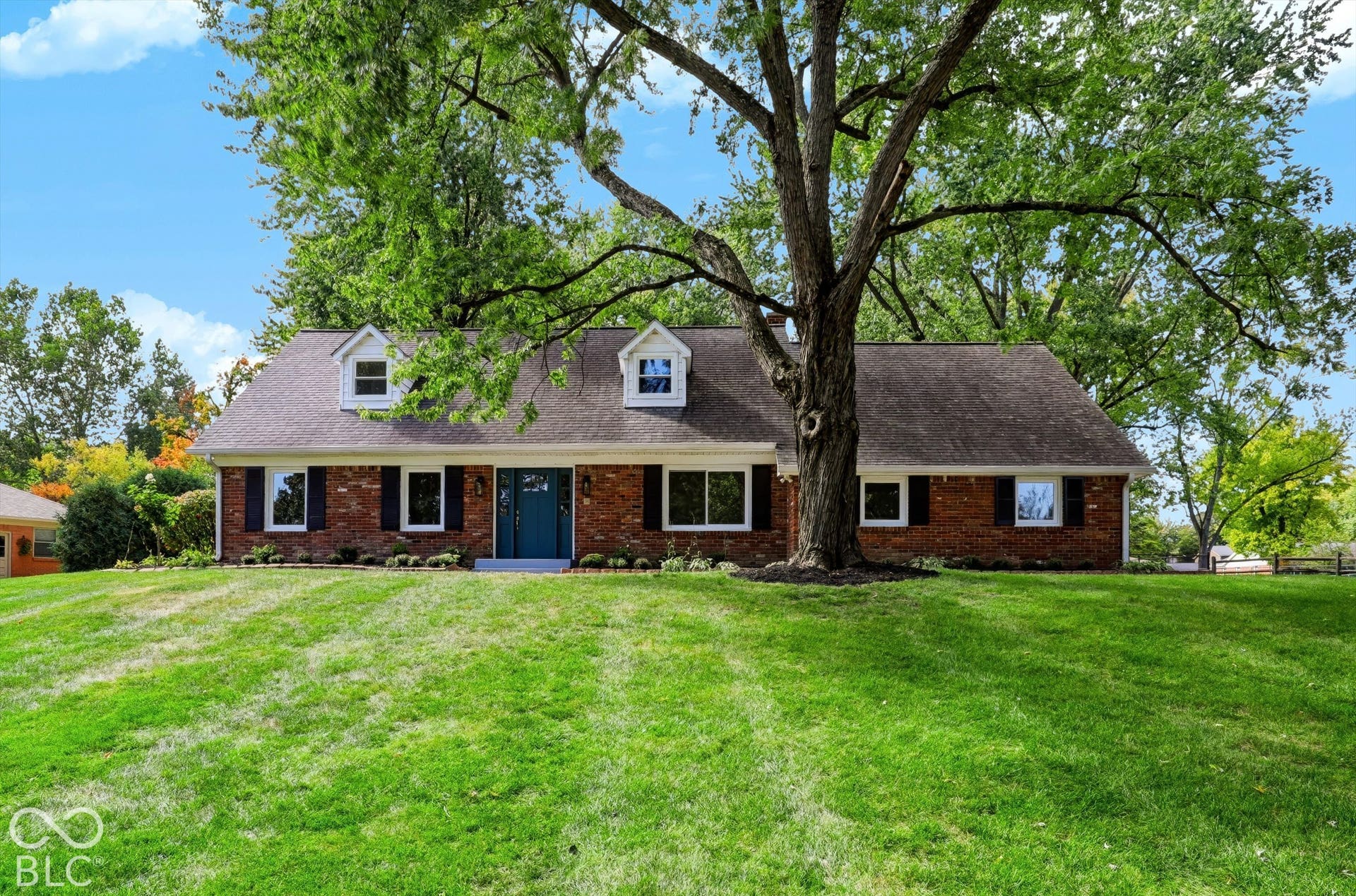Traditional brick exterior featuring a multi-gabled roofline, dormer windows, contrasting shutters, and a prominent mature shade tree