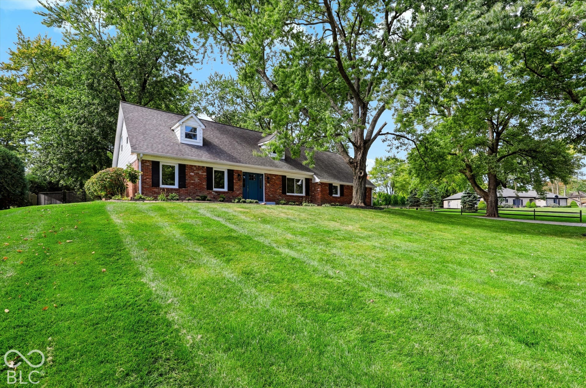 Exterior featuring a brick facade, gabled roofline with dormer windows, expansive manicured lawn, and mature canopy trees