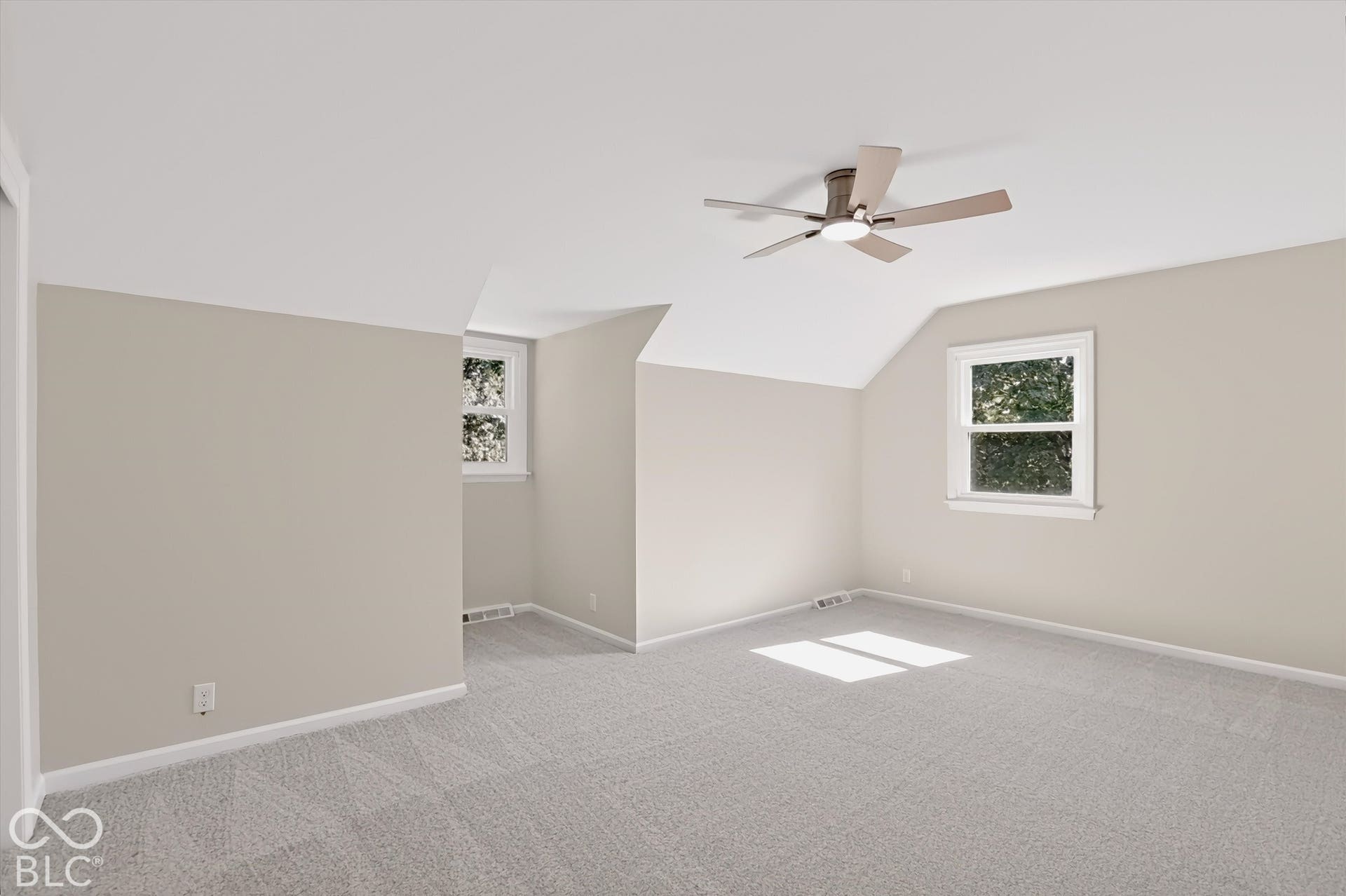 Neutral-toned interior featuring angled ceilings, carpeted flooring, two double-hung windows, and a multi-blade ceiling fan with integrated lighting