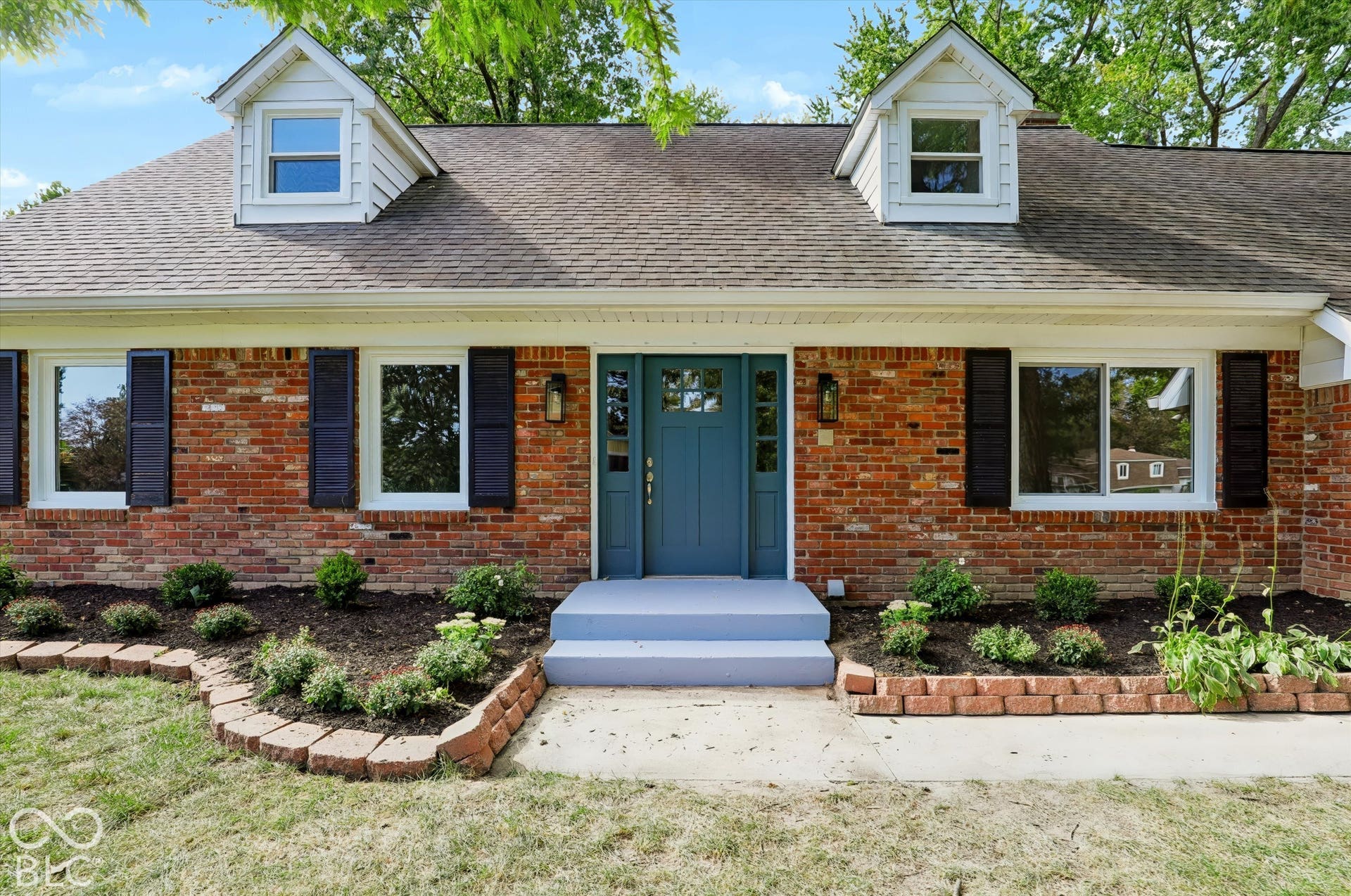 Red brick exterior featuring dual gabled dormers, dark decorative shutters, and a central entryway with sidelights