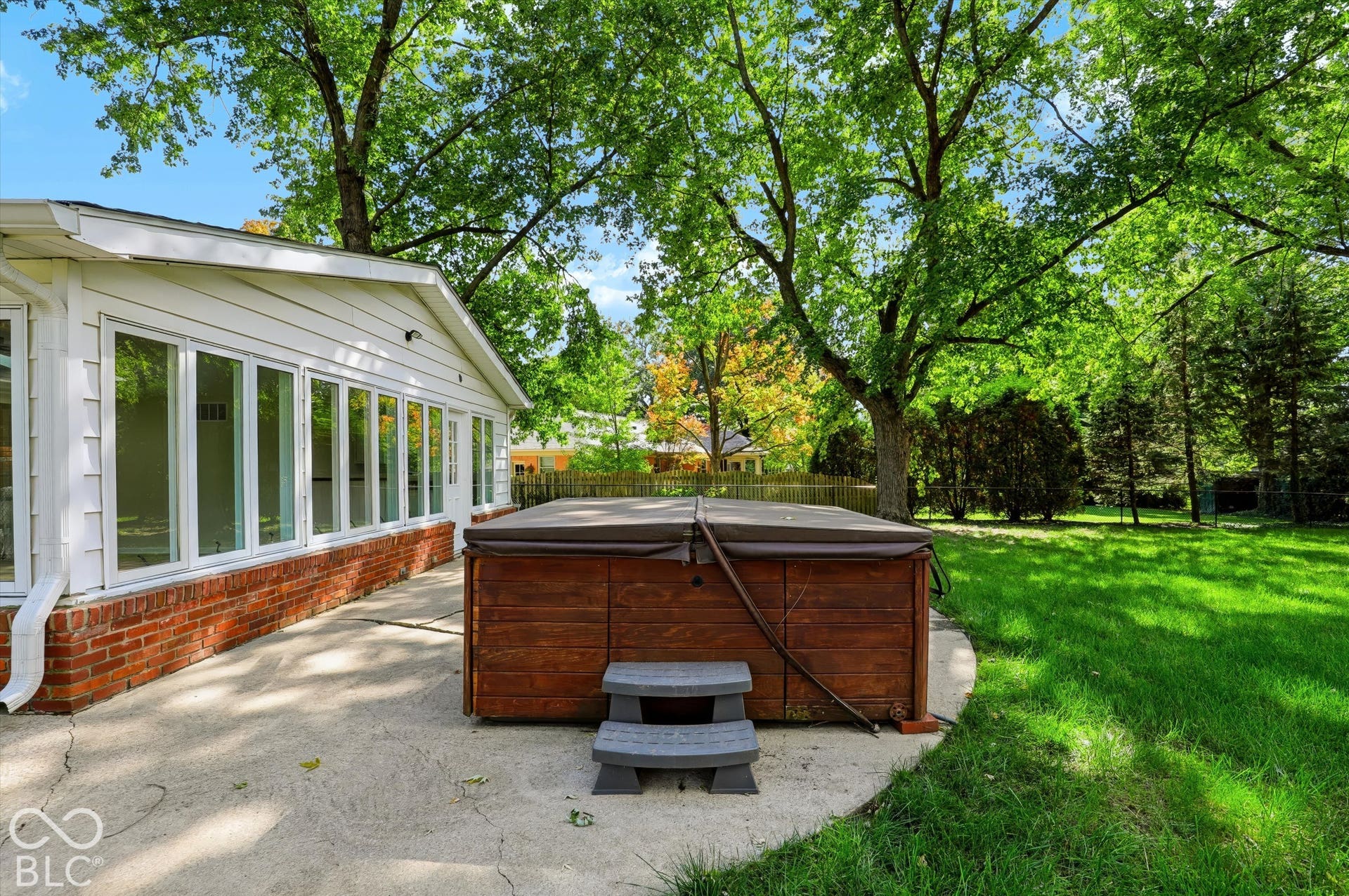 Rear exterior featuring a concrete patio, built-in hot tub, brick and siding facade, and large windows set against a mature treed landscape