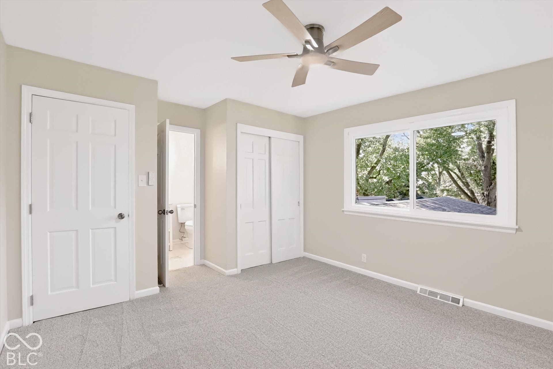 Neutral-toned room featuring neutral carpeting, a multi-blade ceiling fan, six-panel closet doors, and a window with exterior tree foliage