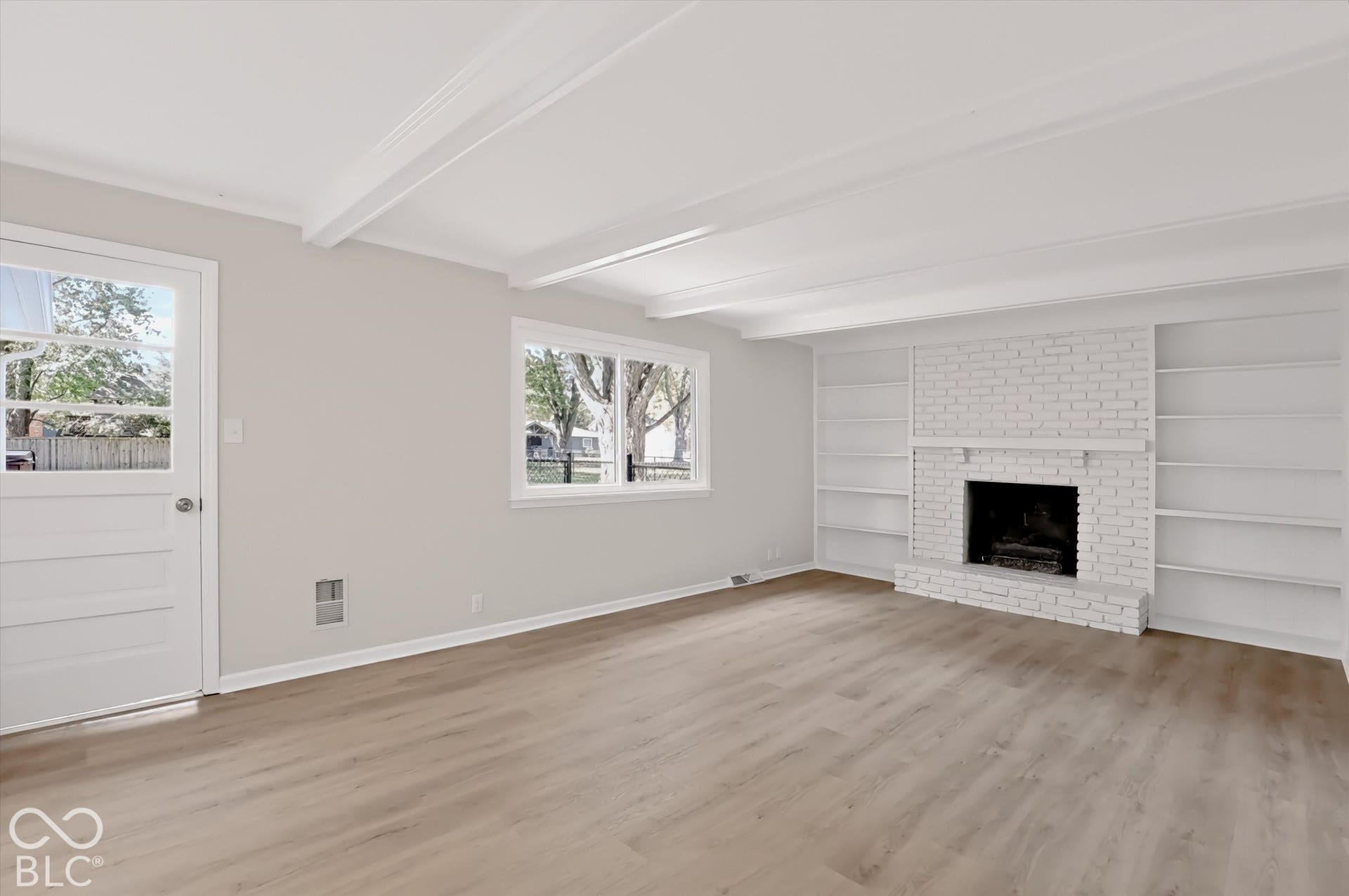 Living area featuring wood-finish flooring, exposed ceiling beams, a painted brick fireplace, and built-in shelving