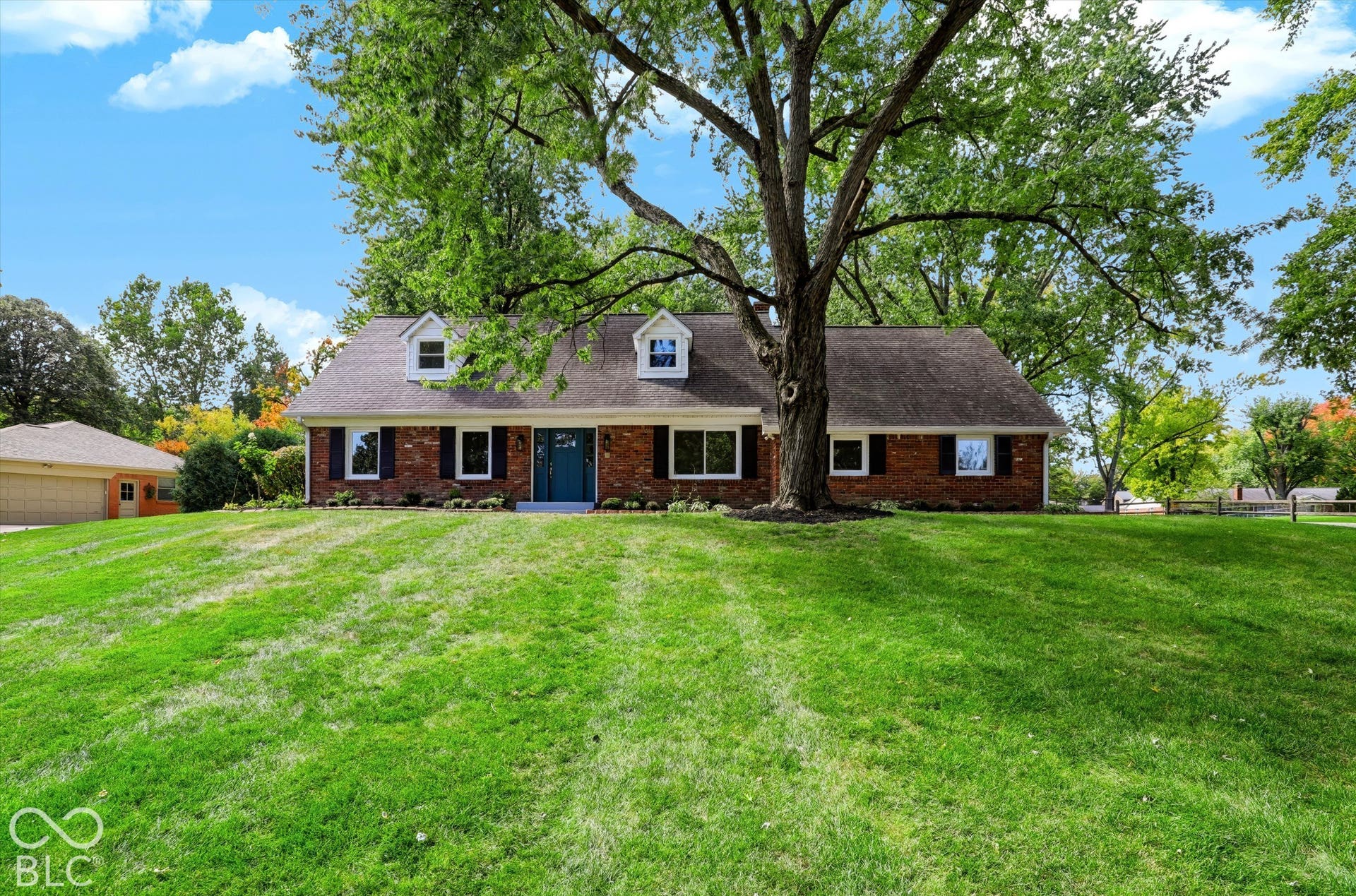 Exterior featuring a brick facade, dormer windows, shingle roofing, and an expansive lawn with mature trees