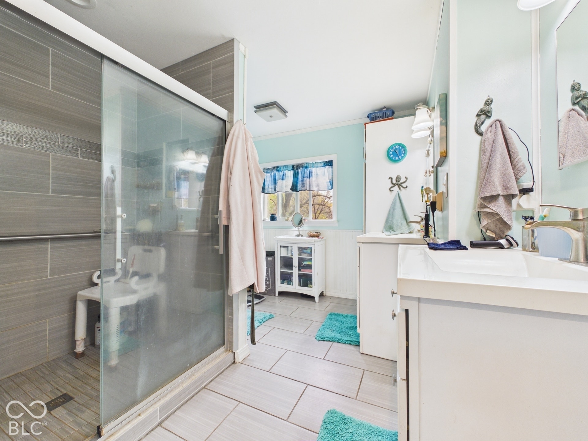 Walk-in shower with floor-to-ceiling gray tile, glass enclosure, and stone-finish flooring, featuring a dual-sink vanity with white cabinetry and metal hardware