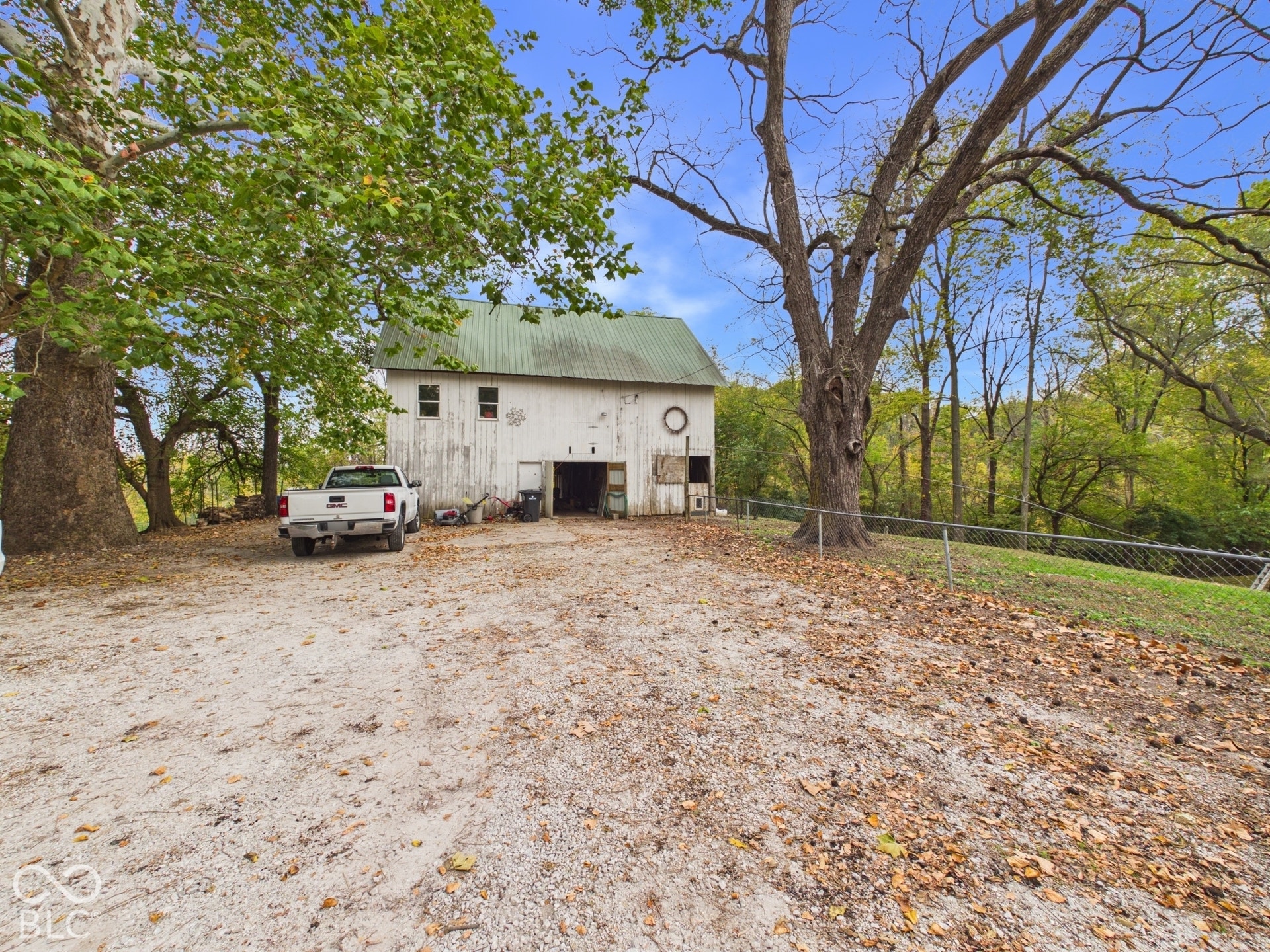 Outbuilding with a green metal roof, light-colored siding, and a gravel lot surrounded by mature trees