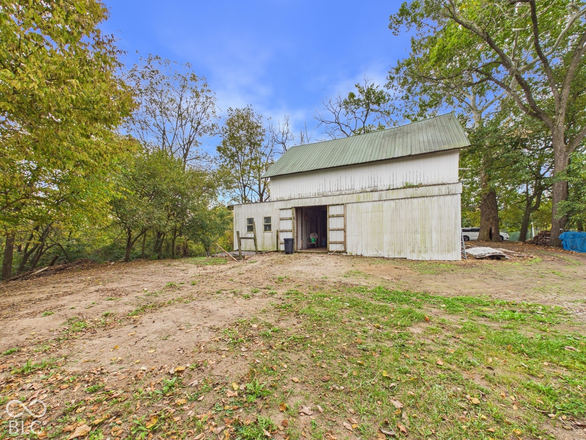 Detached outbuilding featuring white vertical siding, a metal roof, and an open entry point, situated on a lot with mature trees