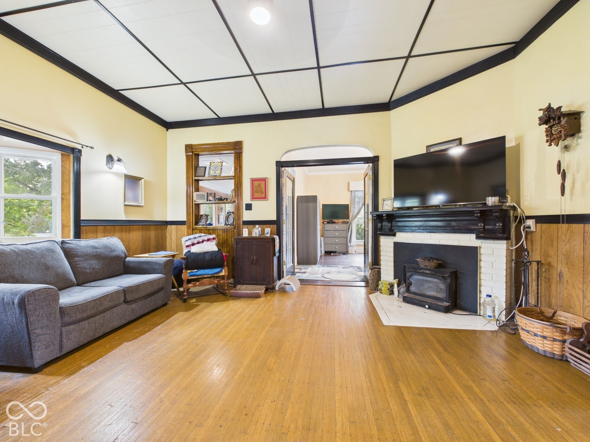 Living area featuring wood-finish flooring, a painted brick fireplace with a mantle, wood wainscoting, built-in shelving, and a coffered ceiling