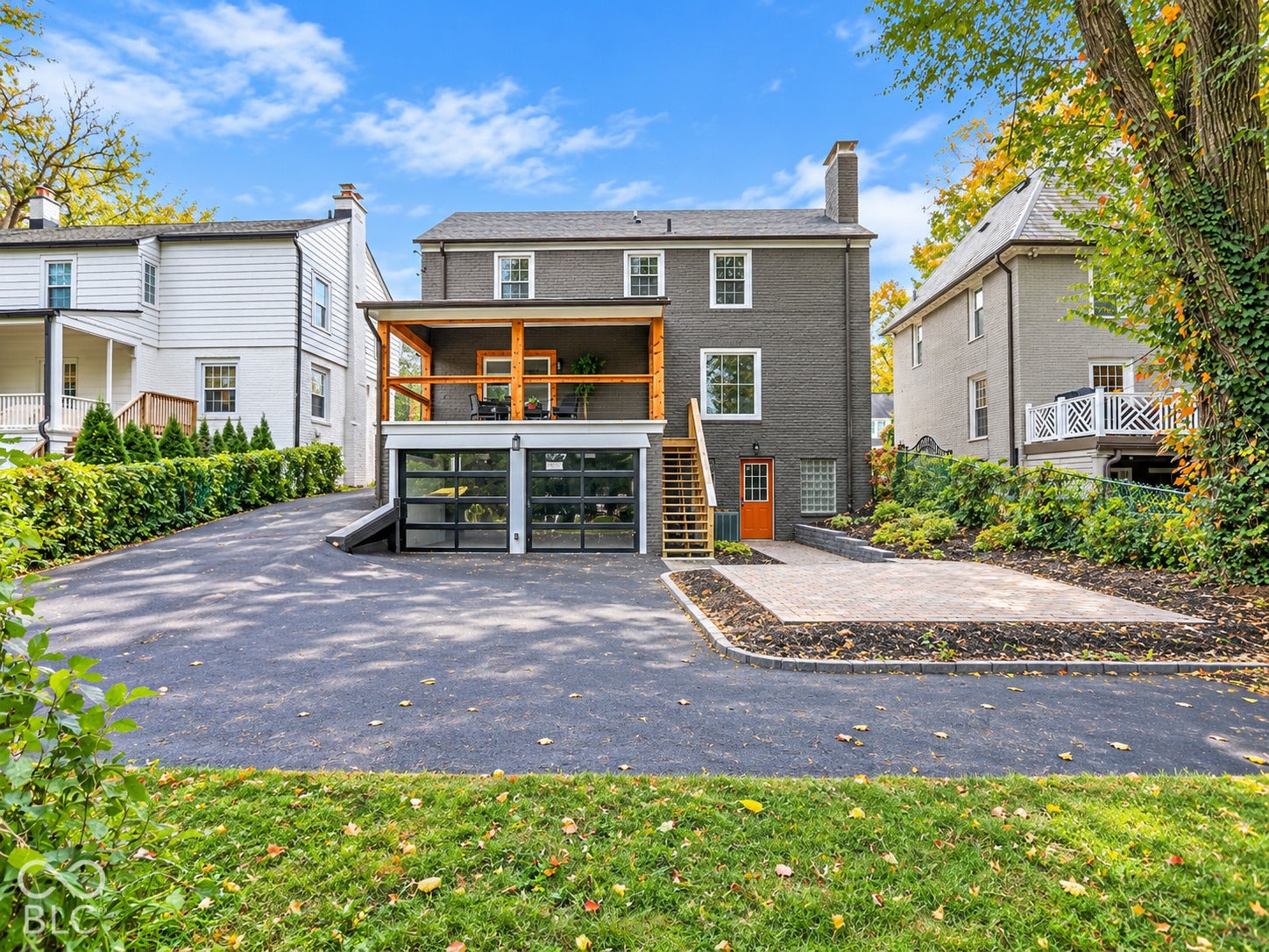 Exterior featuring a dark masonry facade, elevated wooden deck, two glass-paneled garage doors, a stone-paved patio, and asphalt driveway