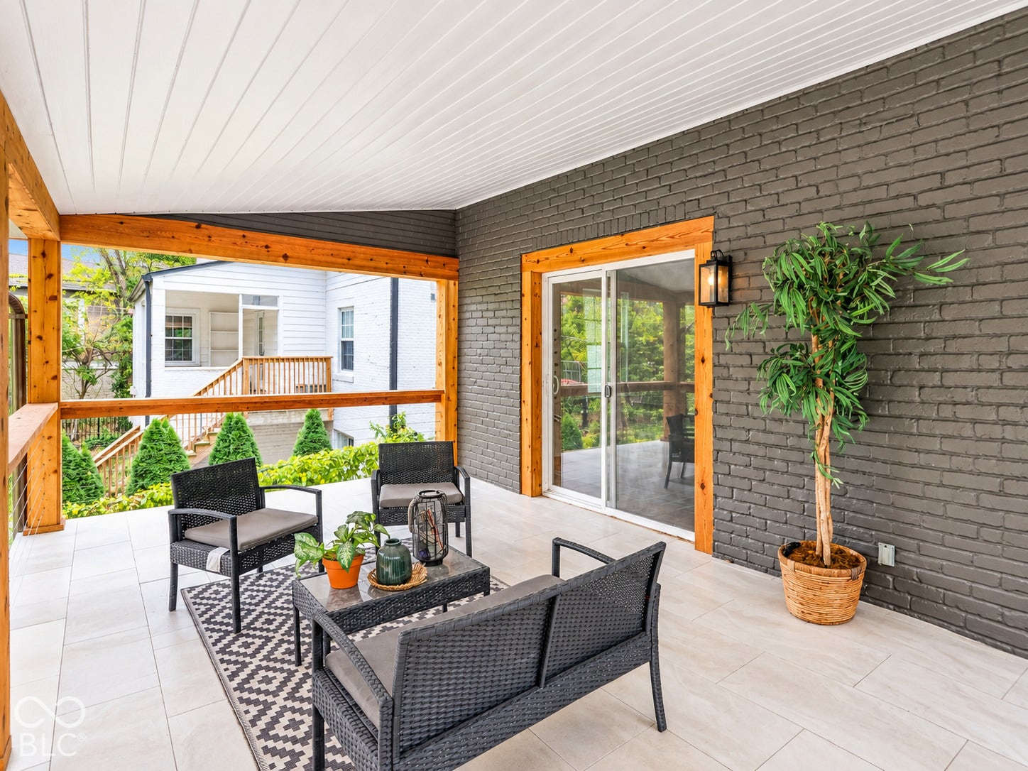 Covered patio featuring large-format porcelain tile flooring, painted brick wall, natural wood-finish exposed structural beams, and sliding glass door entry