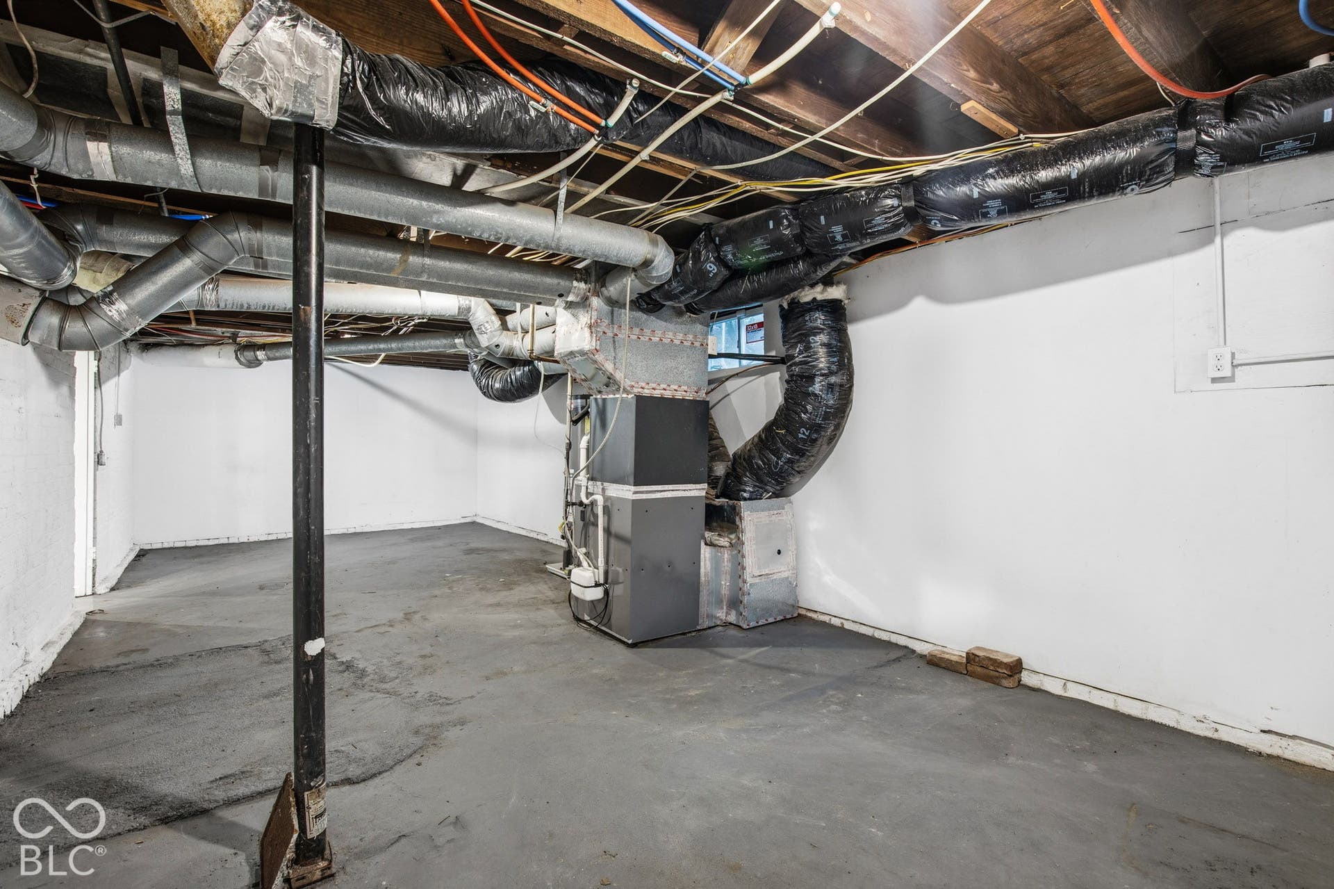 Unfinished lower-level space featuring painted concrete flooring, exposed wooden ceiling joists, and a central forced-air furnace