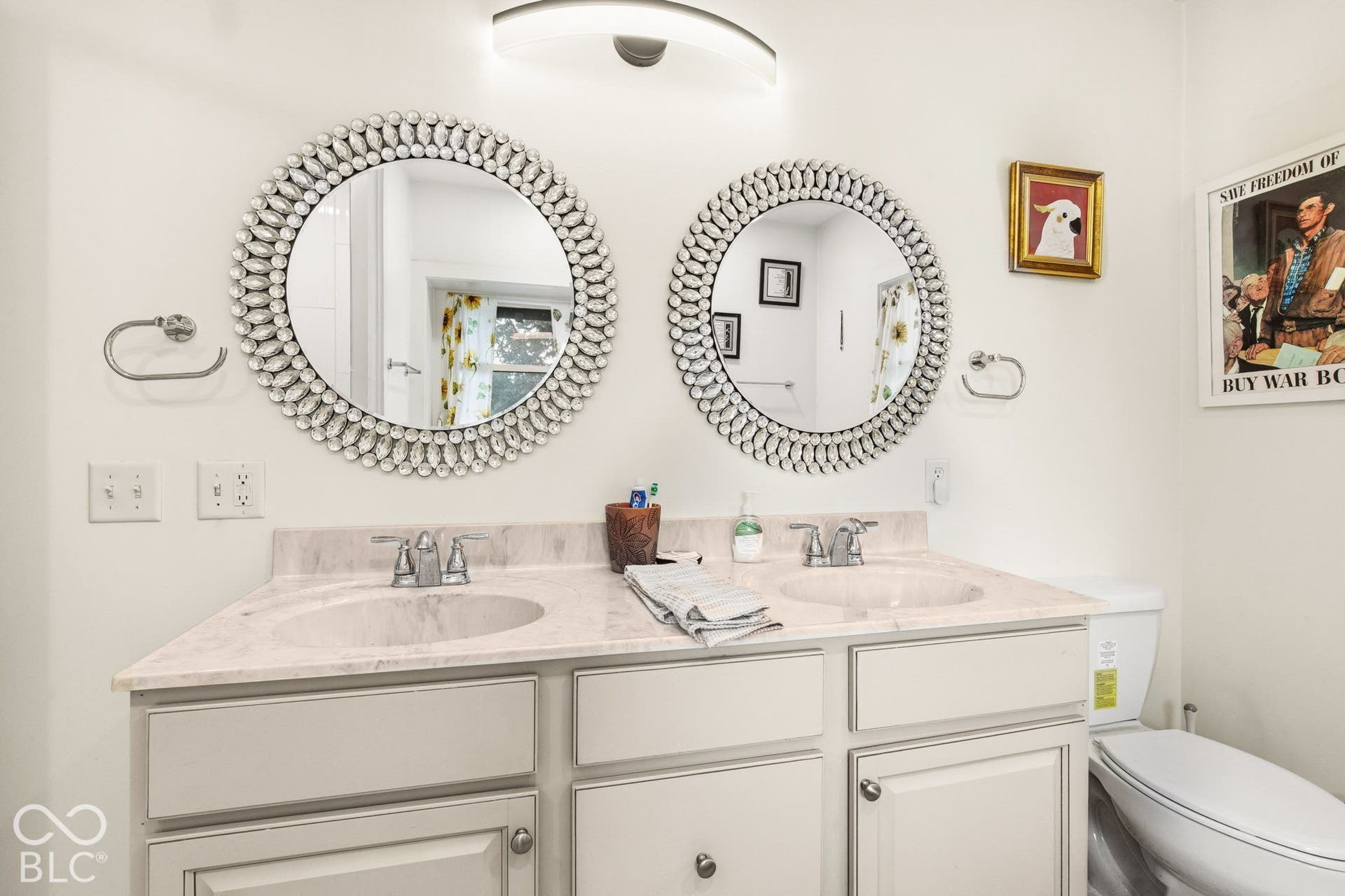 Dual-basin vanity featuring light-toned cabinetry, stone-composite countertop with integrated sinks, dual chrome faucets, and circular decorative mirrors with a curved overhead wall-mounted light fixture