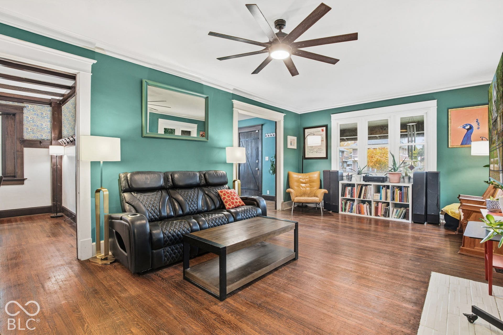 Living area featuring wood-finish flooring, multi-pane windows, a dark-finish multi-blade ceiling fan, and traditional thick-cased door trim