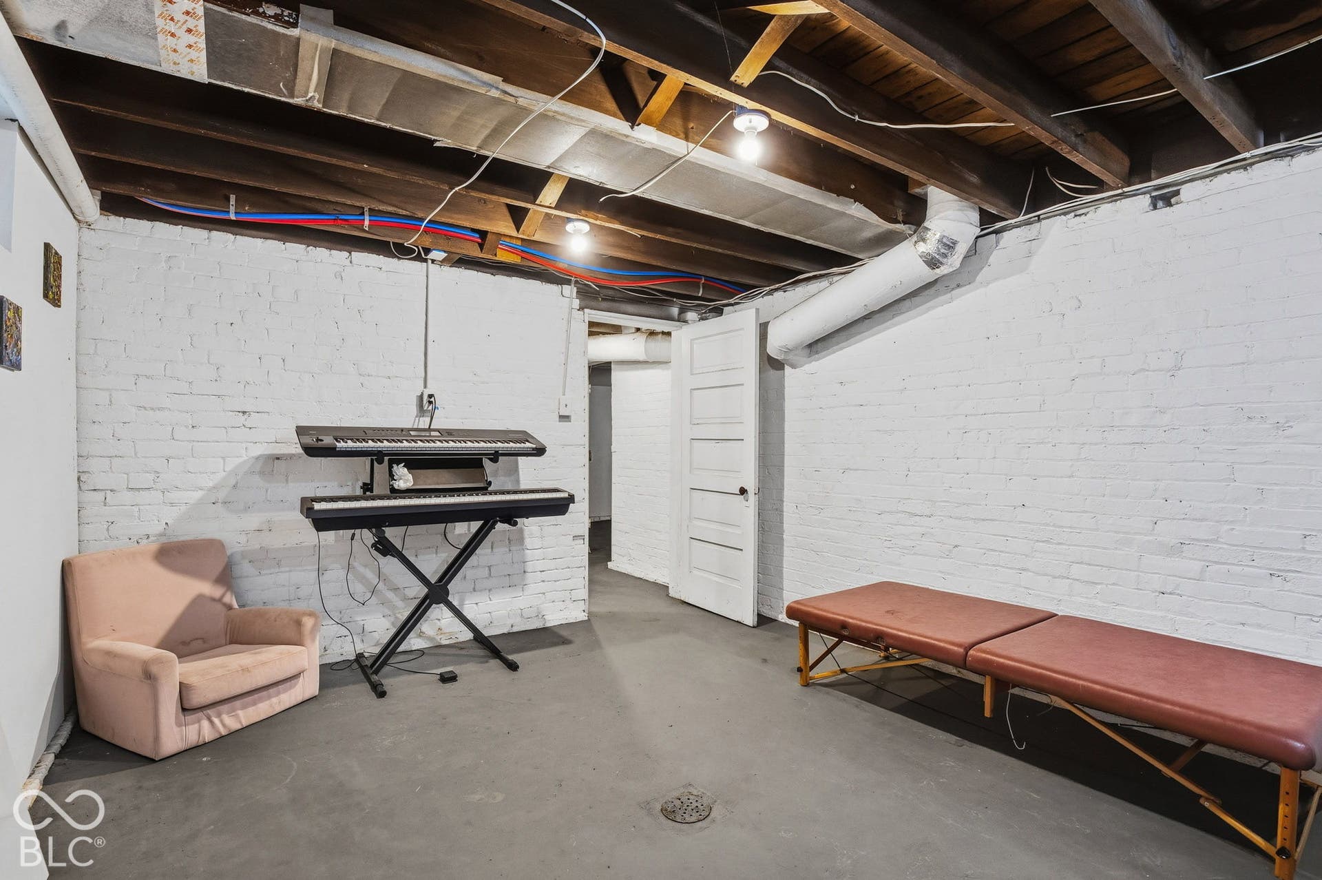 Unfinished lower-level space featuring exposed wood ceiling joists, painted white brick walls, a solid wood paneled door, and concrete flooring with a central floor drain