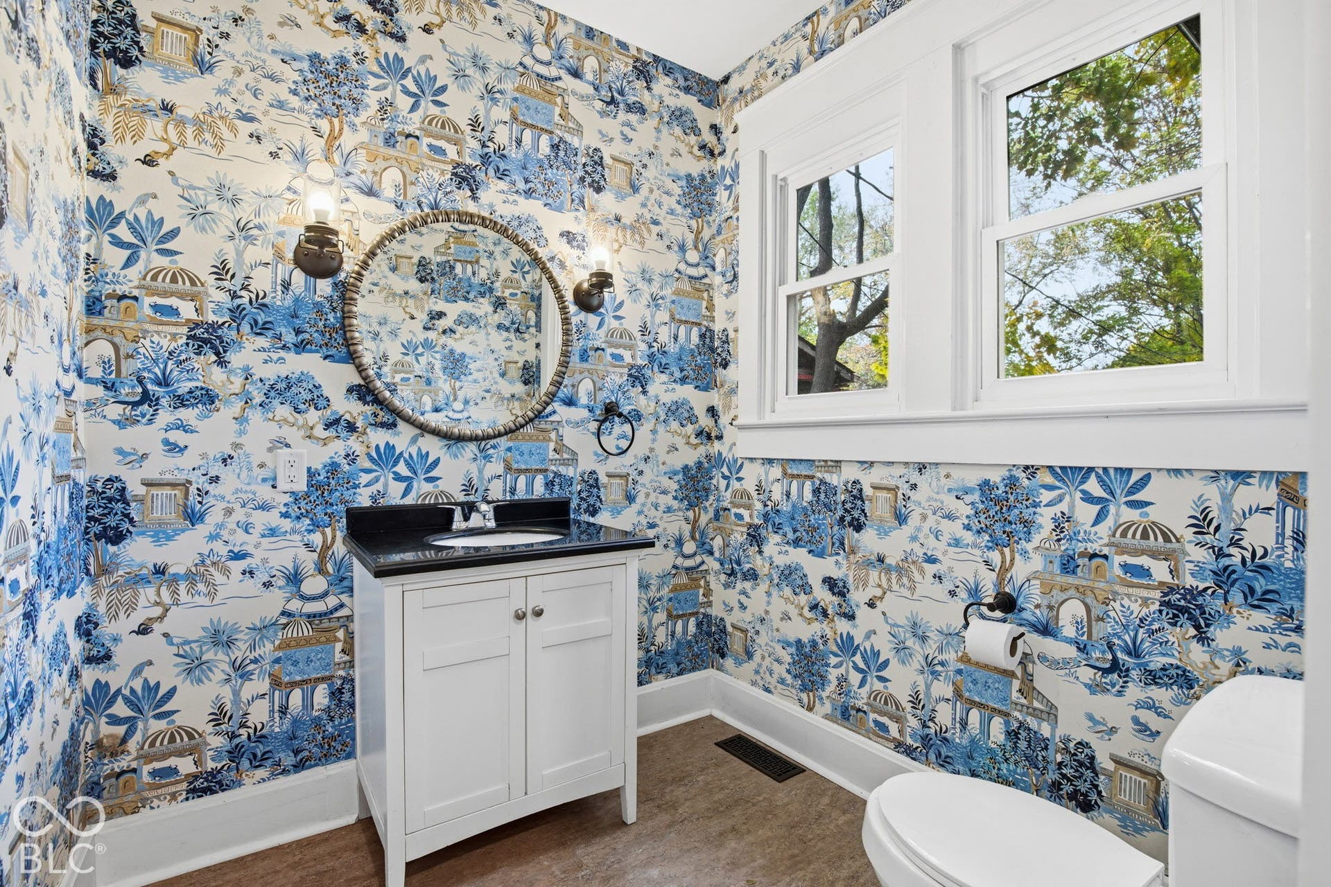 Bathroom featuring a white vanity cabinet, dark stone countertop with integrated sink, circular wall-mounted mirror, dual metallic sconces, and double-hung windows