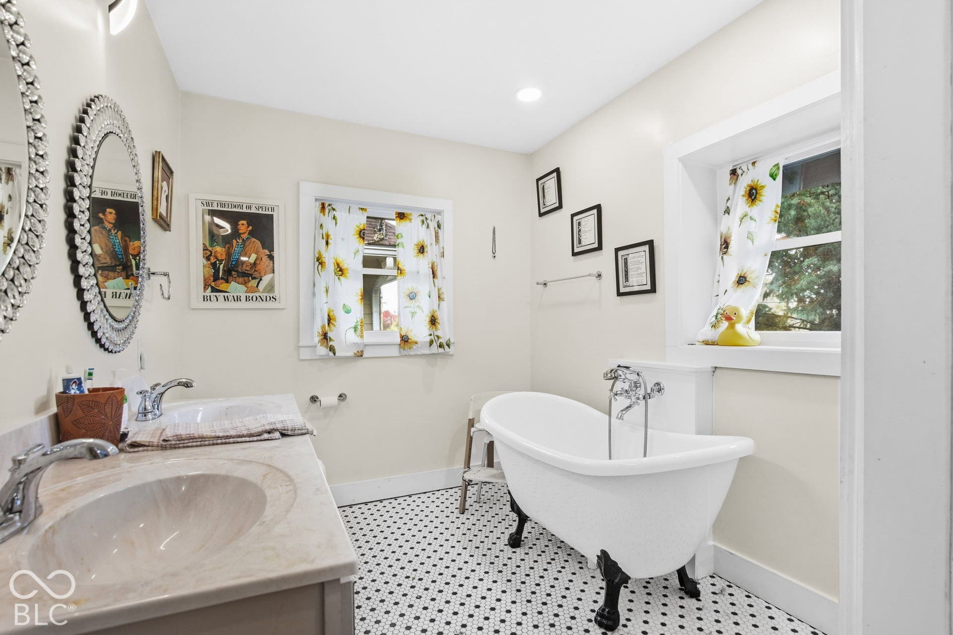 Double vanity with integrated sink basins and stone-finish countertop, white clawfoot soaking tub with polished chrome hardware, and black-and-white hexagonal tile flooring