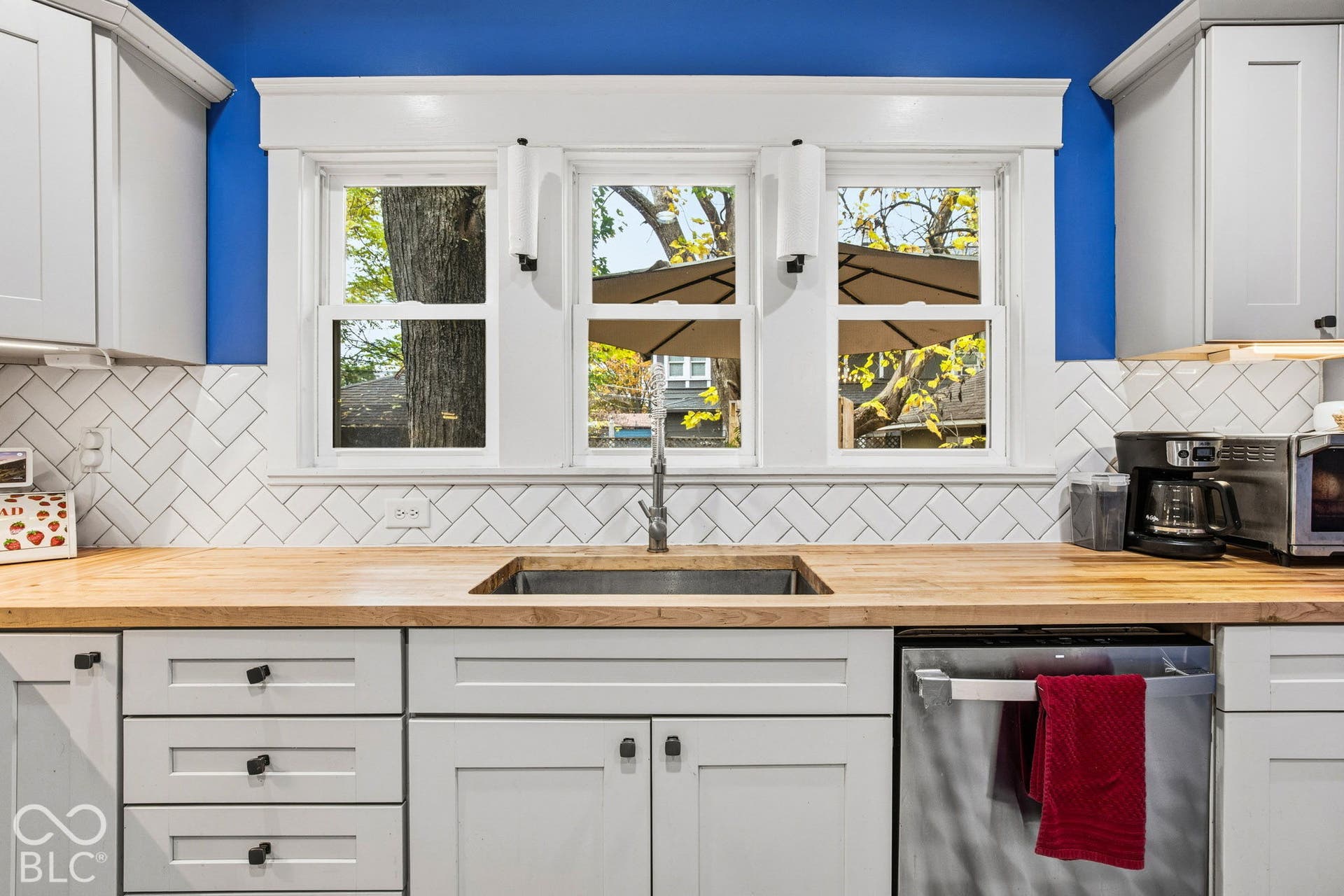 Light gray shaker-style cabinetry, butcher block countertops, white herringbone tile backsplash, stainless steel undermount sink, and white-trimmed triple-pane window