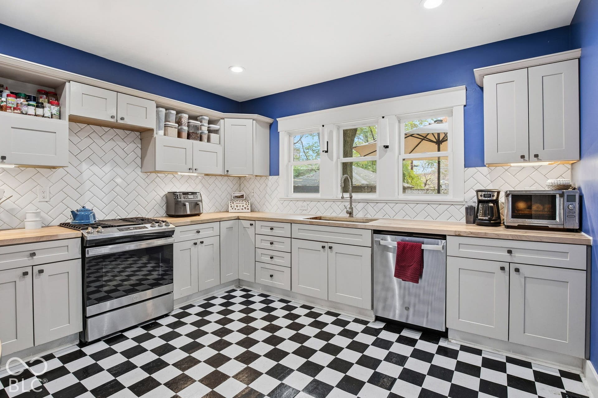 Kitchen featuring light-gray shaker-style cabinetry, butcher block countertops, white herringbone tile backsplash, stainless steel appliances, and checkered tile flooring