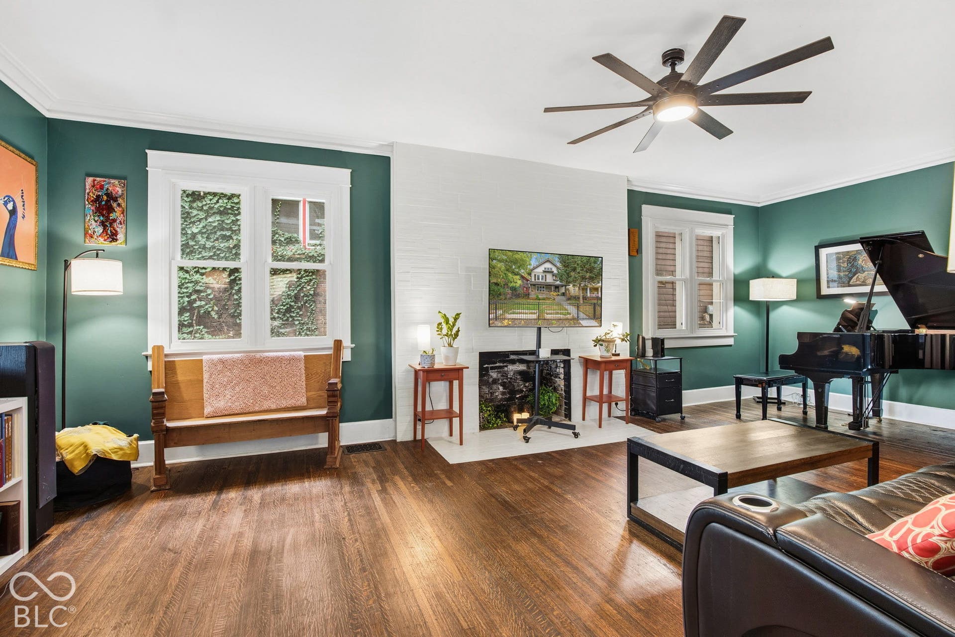 Living area featuring wood-finish flooring, white-painted brick fireplace surround, multi-pane windows with thick trim, and an oversized ceiling fan with integrated lighting