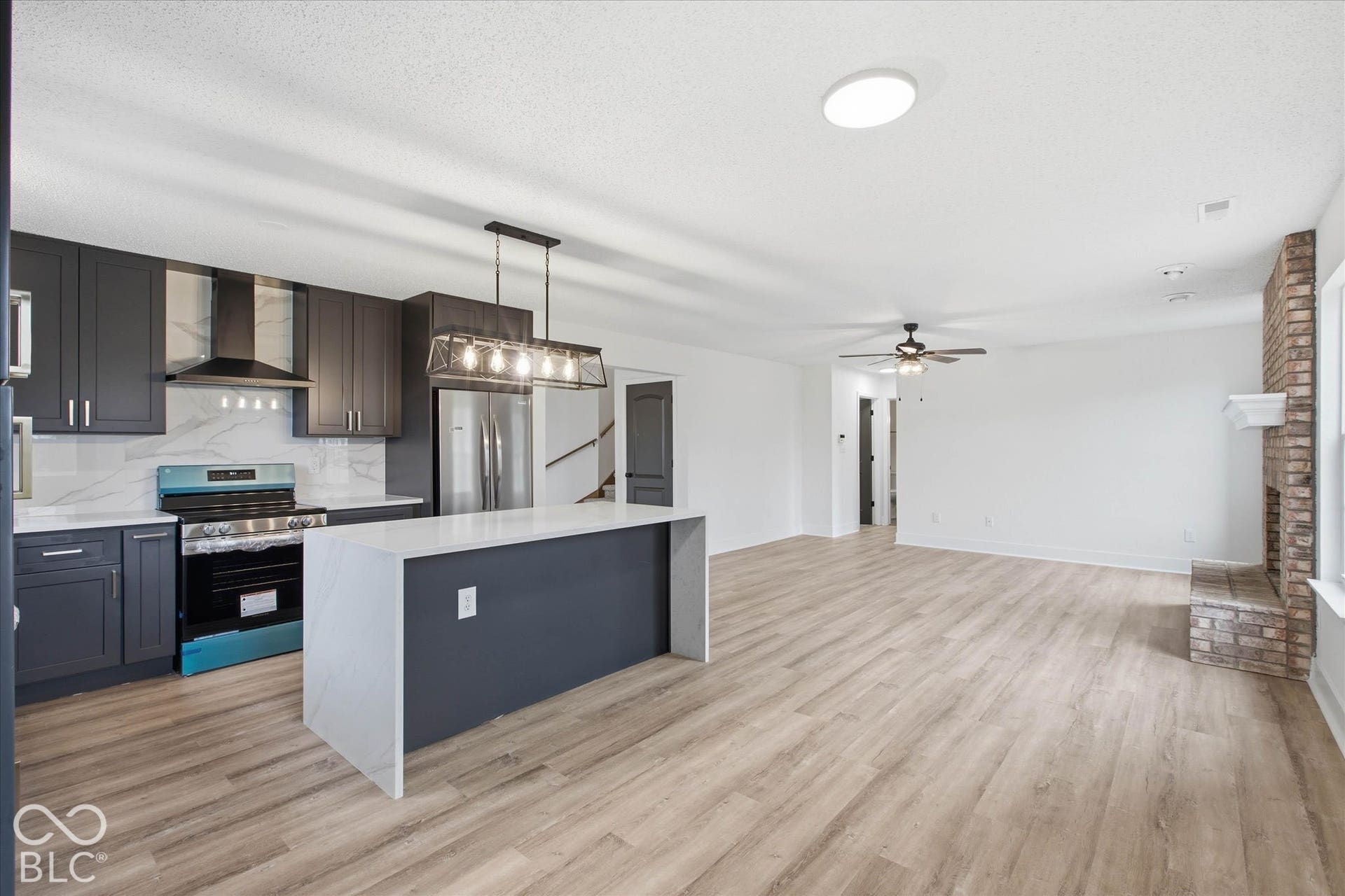 9607 Piper Lake Drive, Indianapolis, IN 46239 Kitchen featuring dark cabinetry, stone-style waterfall island, stainless steel range with overhead hood, wood-finish flooring, and a brick fireplace