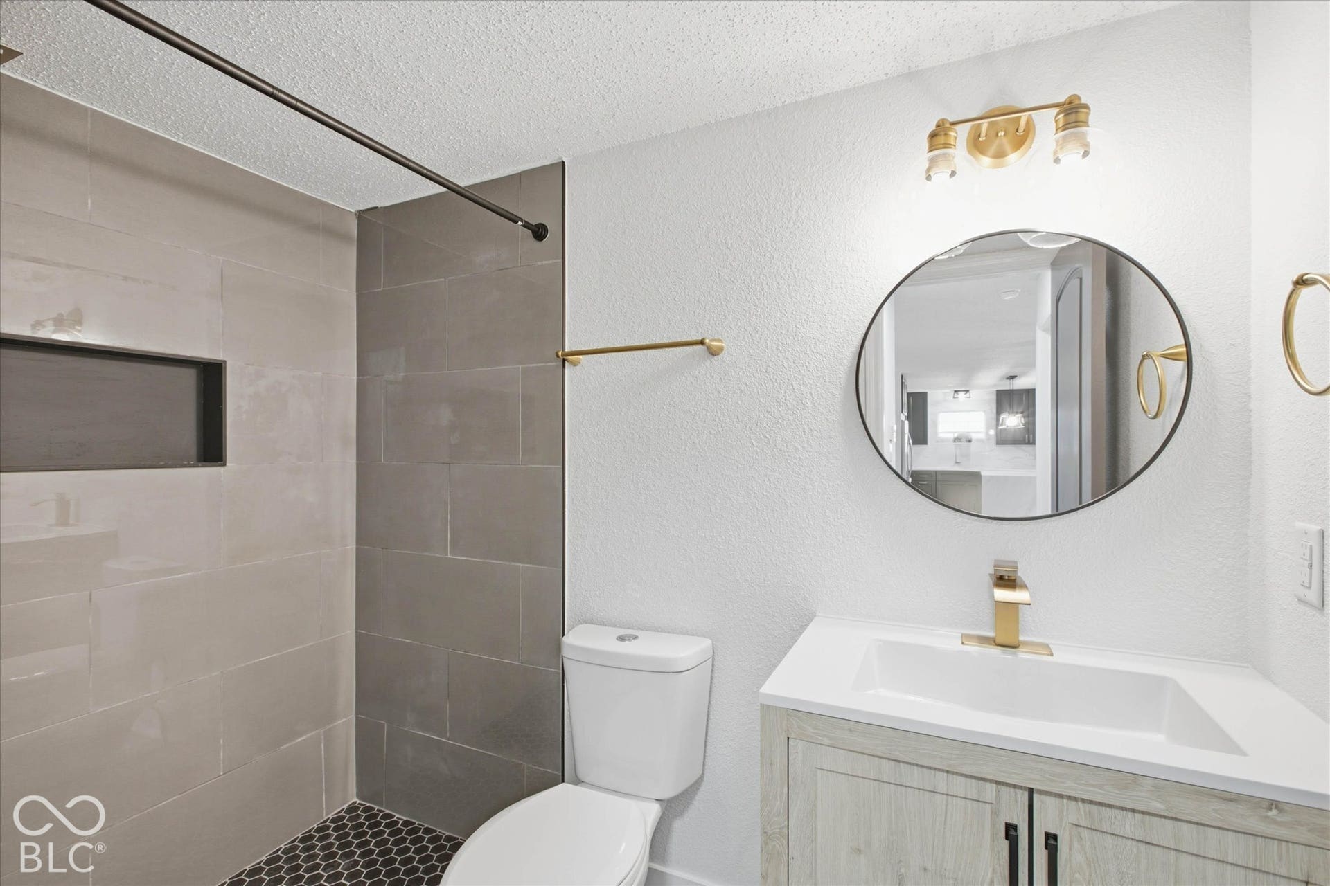 9607 Piper Lake Drive, Indianapolis, IN 46239 Bathroom featuring a wood-finish vanity with a white countertop, circular mirror, metallic wall-mounted lighting, and a shower enclosure with large-format wall tile, built-in shelving, and dark hexagonal floor tile