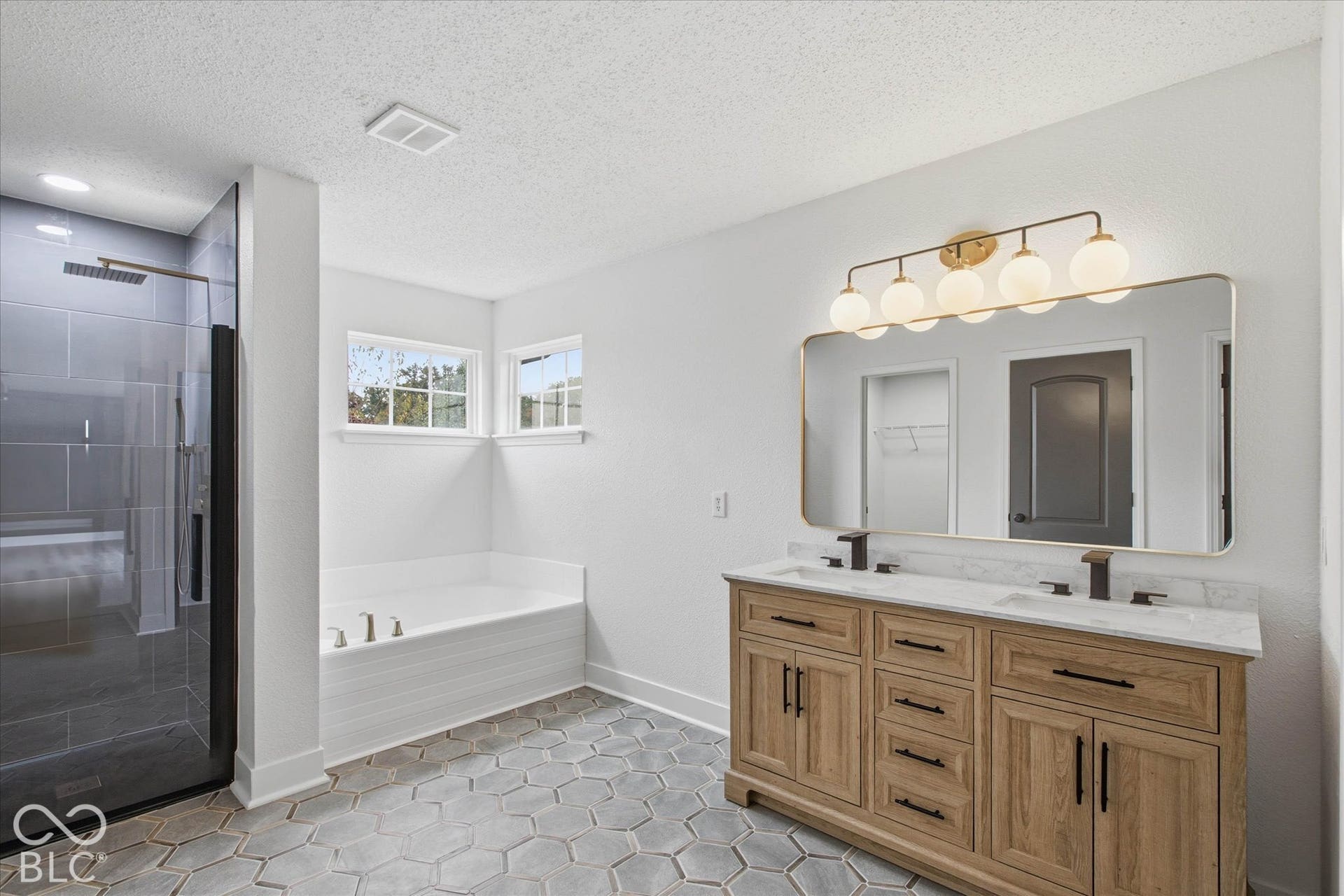 9607 Piper Lake Drive, Indianapolis, IN 46239 Dual-sink vanity with light wood cabinetry, stone-finish countertop, and matte black fixtures, paired with hexagonal tile flooring, a soaking tub, and a glass-enclosed walk-in shower