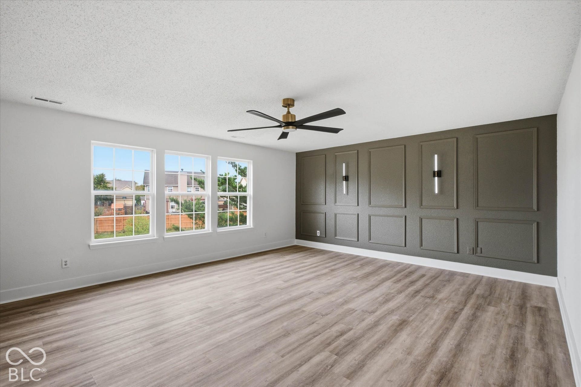 9607 Piper Lake Drive, Indianapolis, IN 46239 Room featuring wood-finish flooring, a charcoal-toned accent wall with decorative molding, dual wall-mounted light fixtures, a multi-blade ceiling fan, and large windows