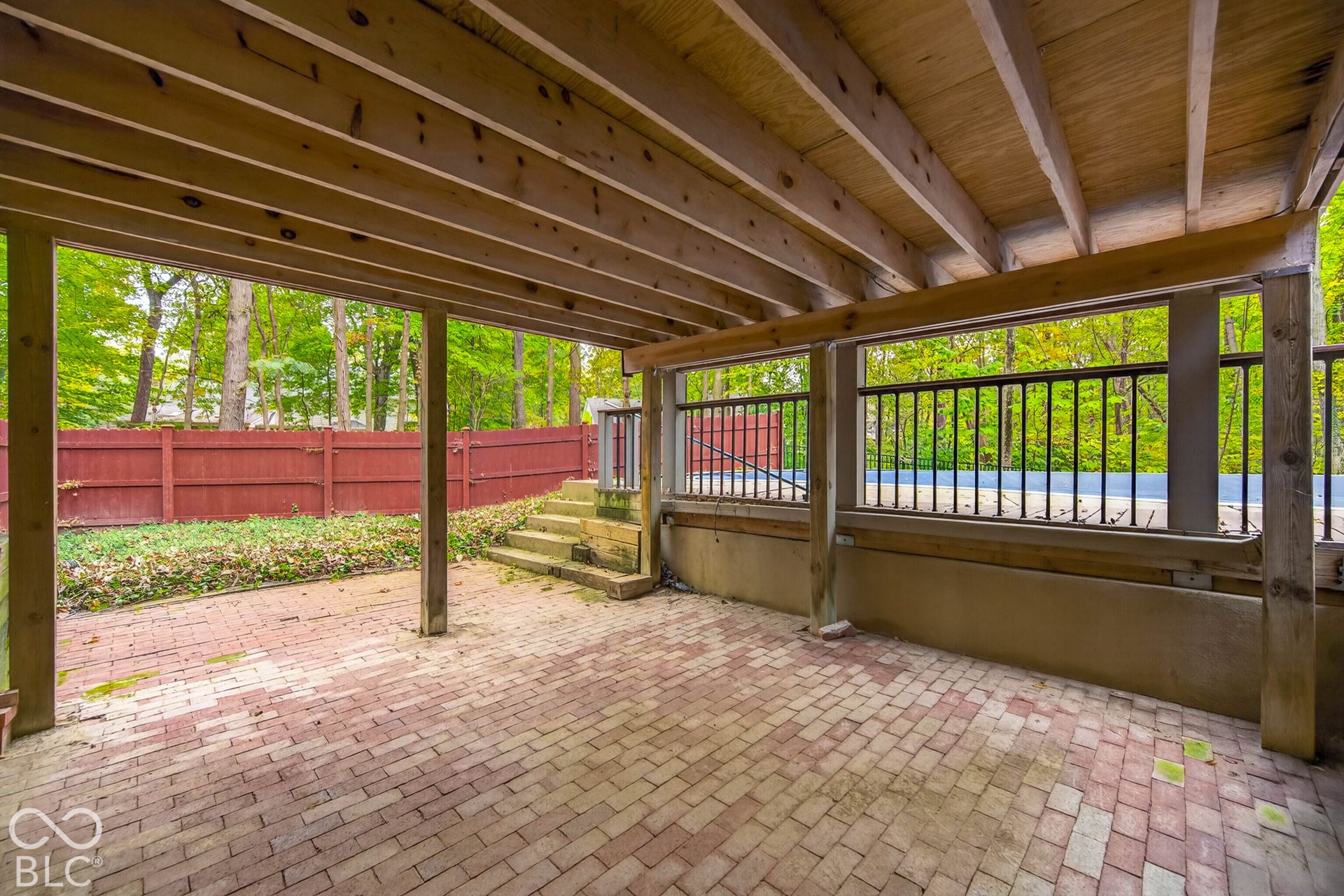 Covered patio featuring red brick pavers, exposed wood ceiling joists, concrete retaining walls, and a black metal railing